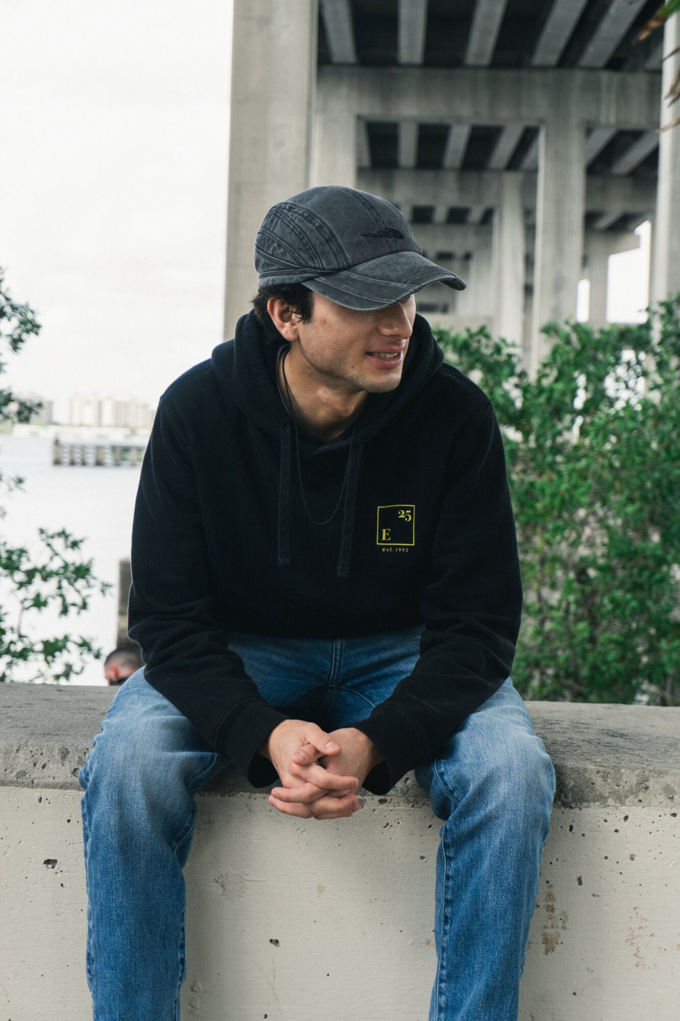 Young man wearing a black cap and hoodie, sitting outdoors with greenery and a bridge structure in background.