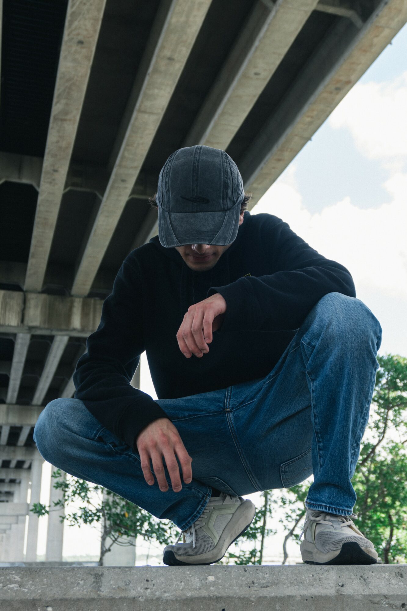 Person crouching under a bridge wearing a cap, black sweater, and jeans, with trees and sky in background.
