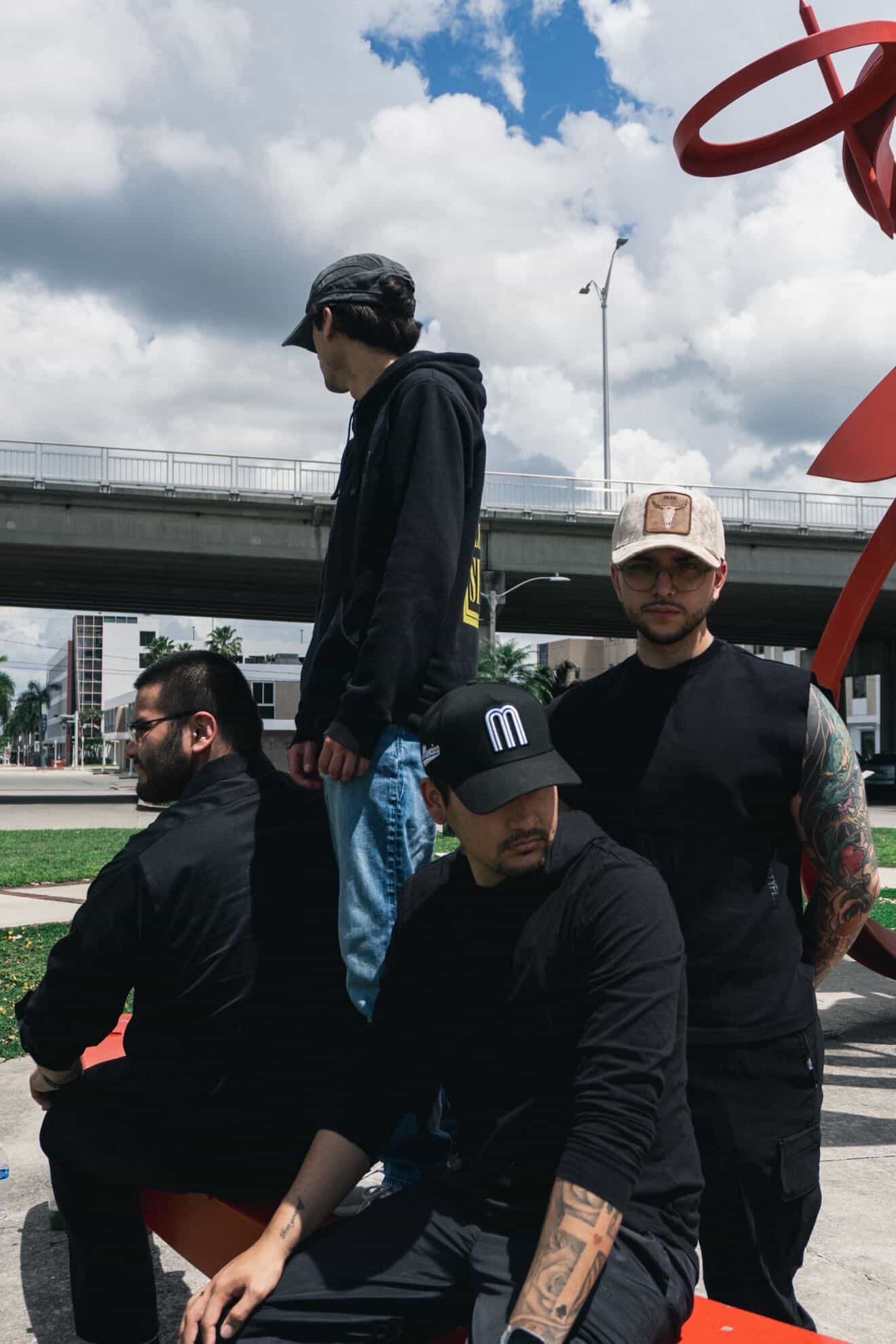 Four men outdoors near a bridge, some sitting and one standing, with cloudy sky and urban background.