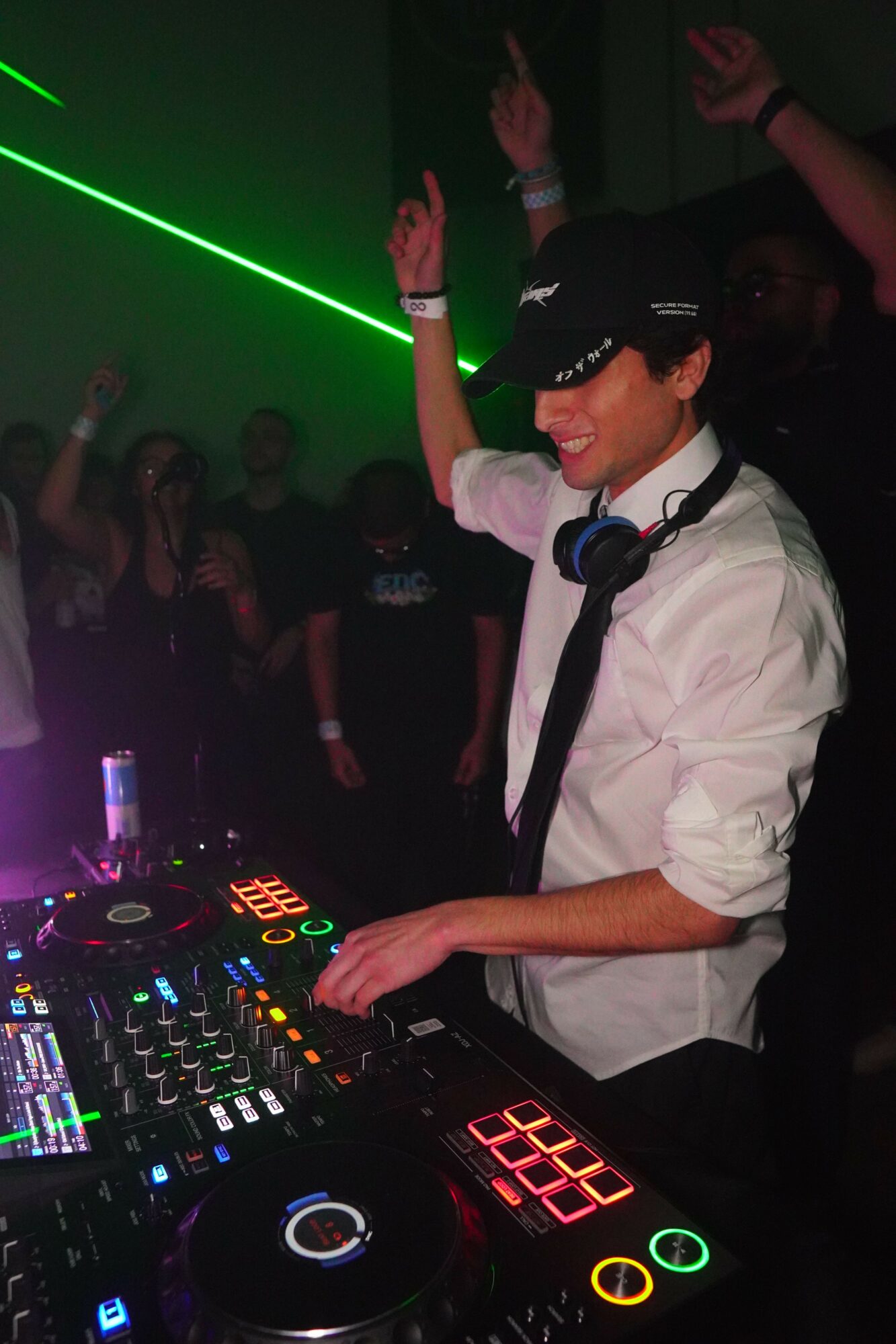 DJ wearing a cap and headphones, smiling, with people dancing and raising hands in a dark club with green laser lights.