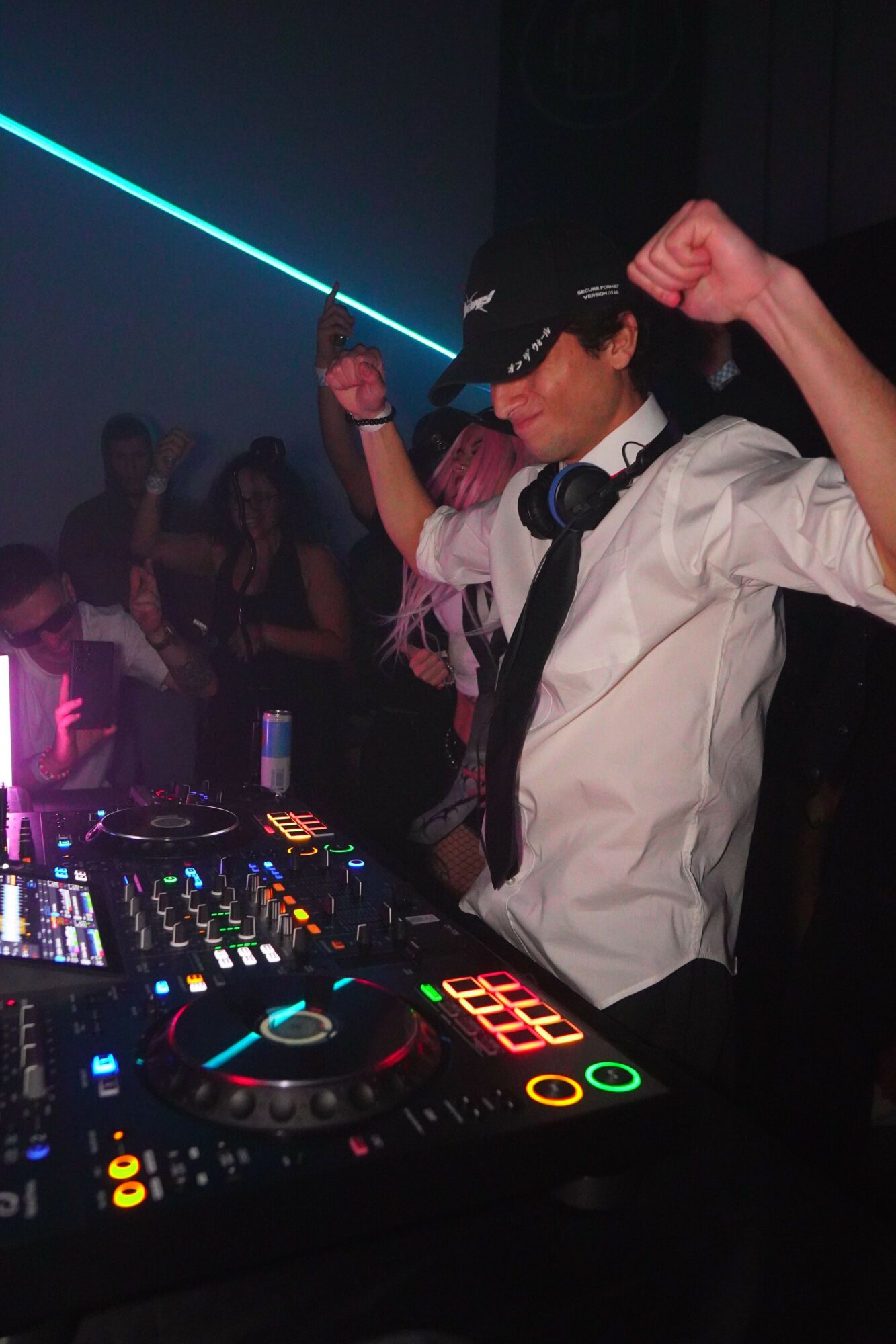 DJ wearing headphones and a cap, raising a fist, with a DJ mixer and people dancing in the background.