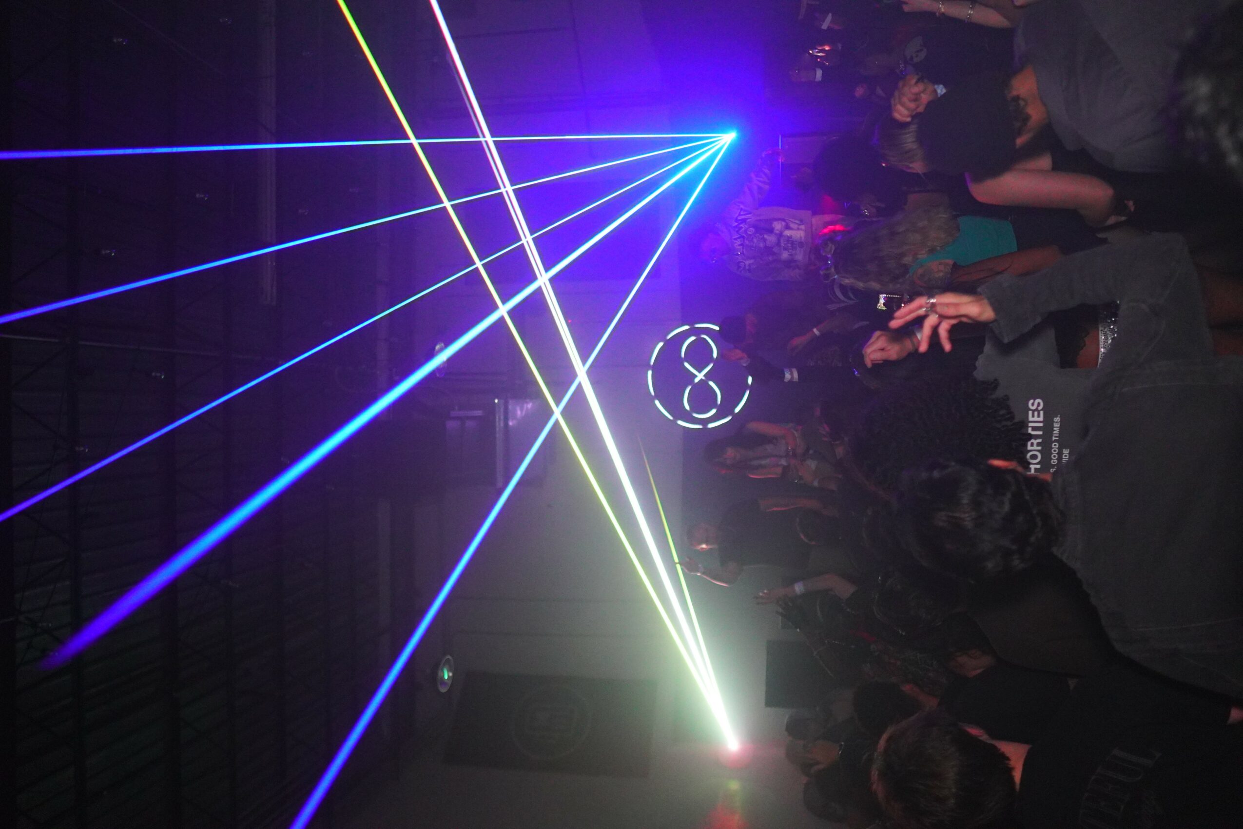 People in a dark room with colorful laser lights and a DJ setup, some dancing or watching the lights.