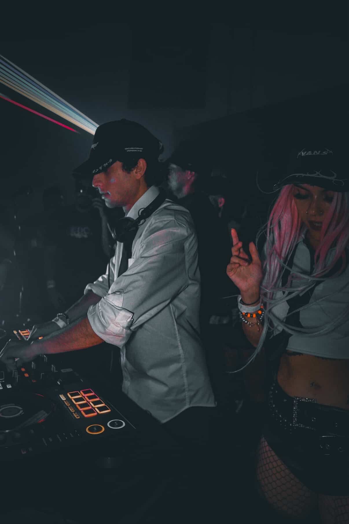 Two people DJing in a dark club with colorful light streaks, one wearing a cap and the other with pink hair.
