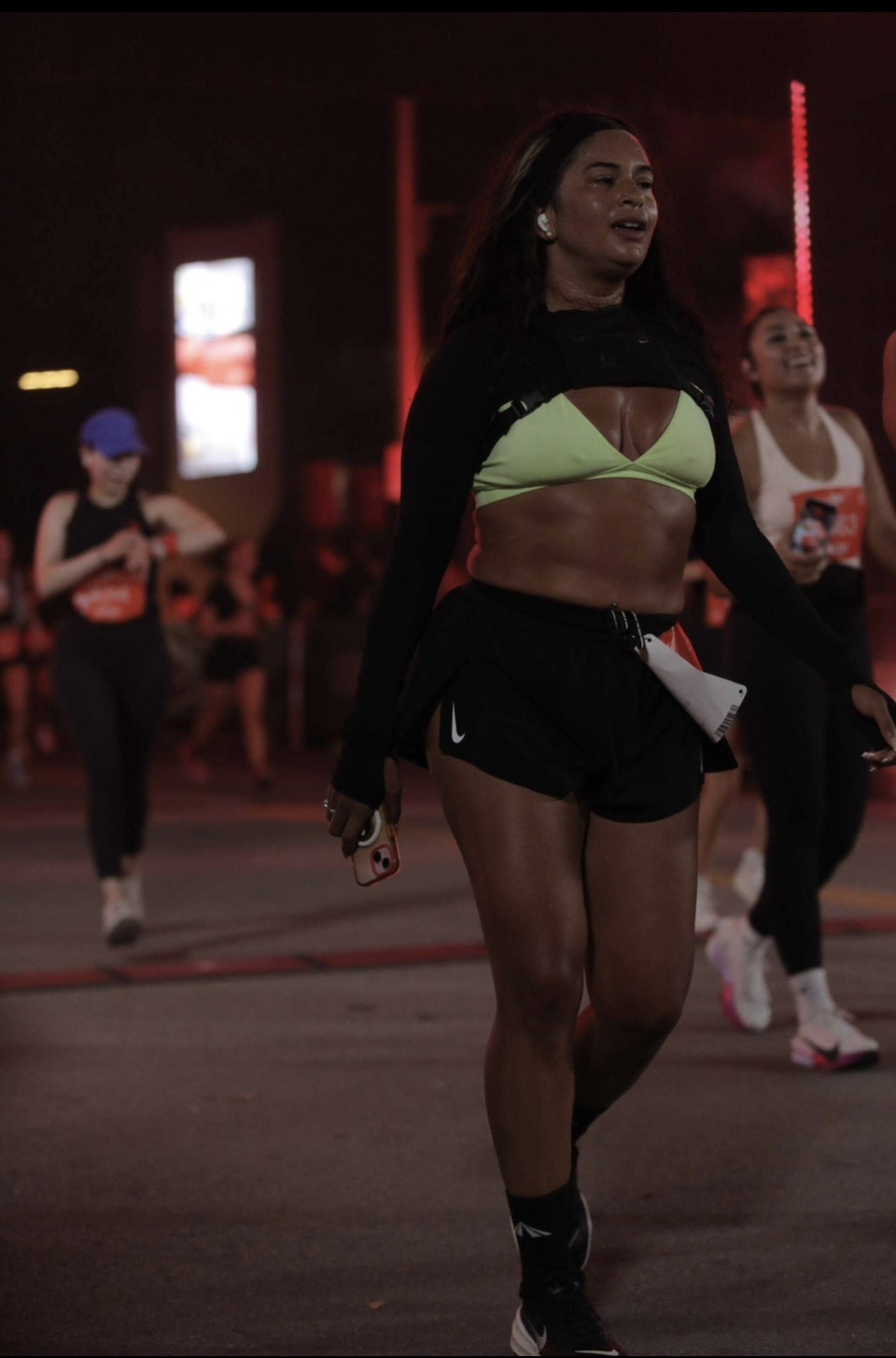 Woman walking outdoors at night, wearing a green sports bra, black shorts, and a black jacket, with other people in background.