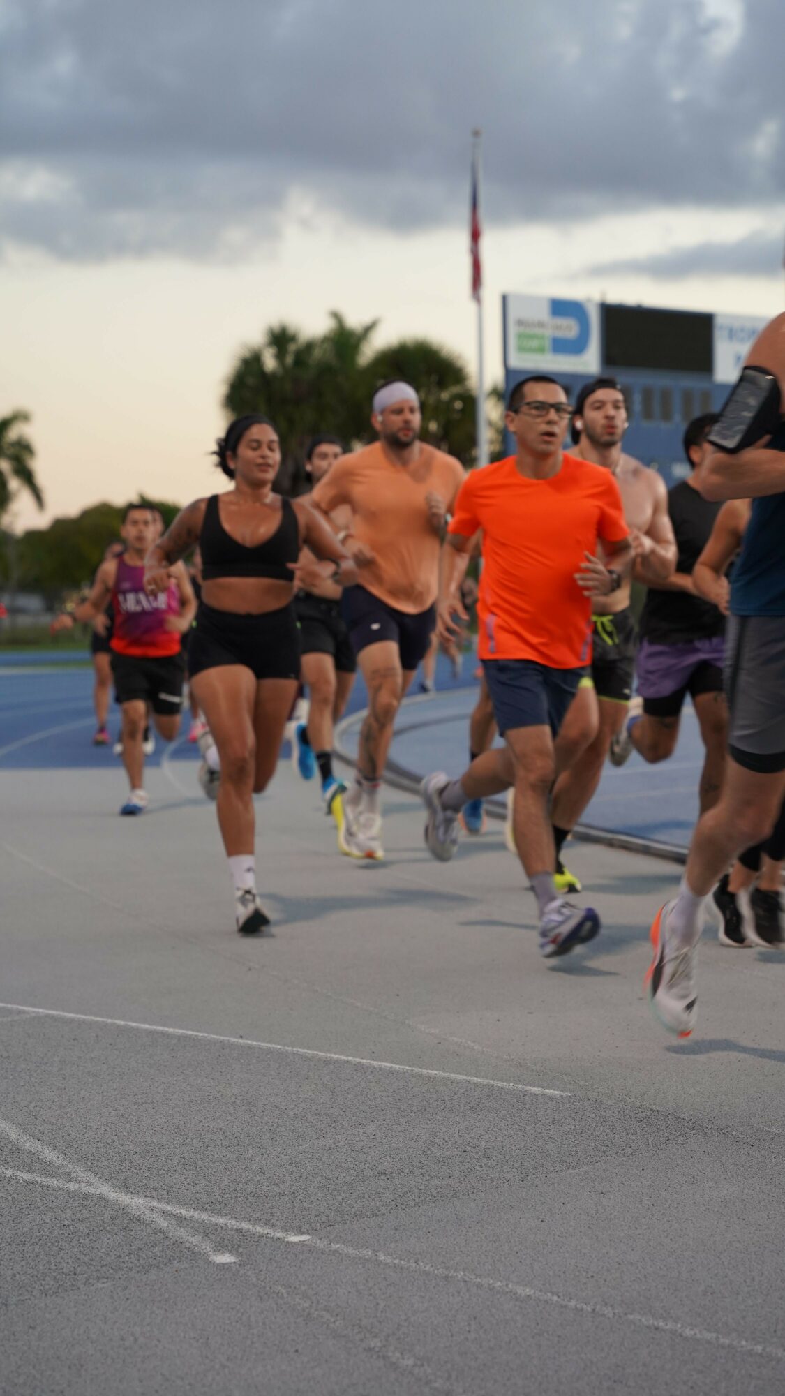 Group of people running on a track outdoors, some wearing athletic clothing, with a cloudy sky and a building in background.