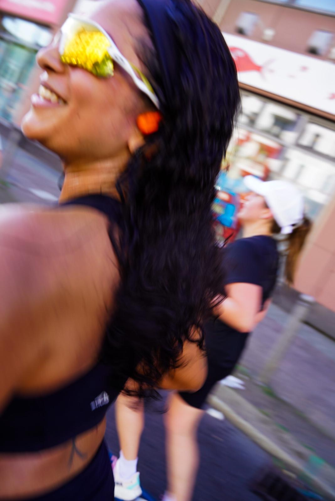 Smiling woman with long dark hair, wearing sunglasses with yellow flowers, outdoors in a busy area.