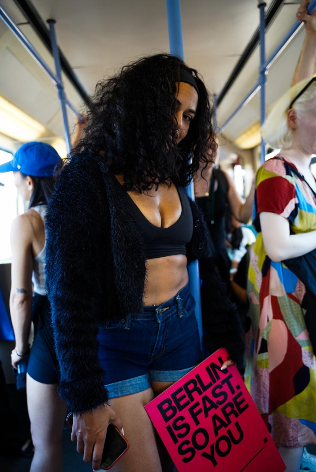 Woman with curly hair wearing a black crop top and shorts, holding a phone, surrounded by people.
