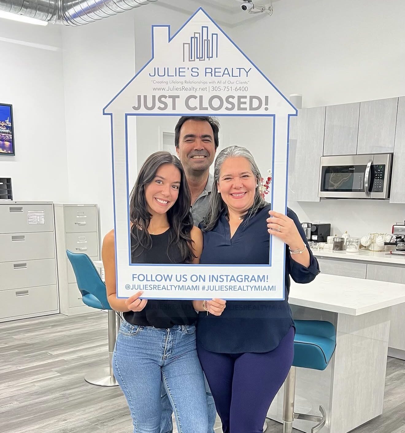 Three people holding a house-shaped sign that says 'Just Closed!' inside an office space.