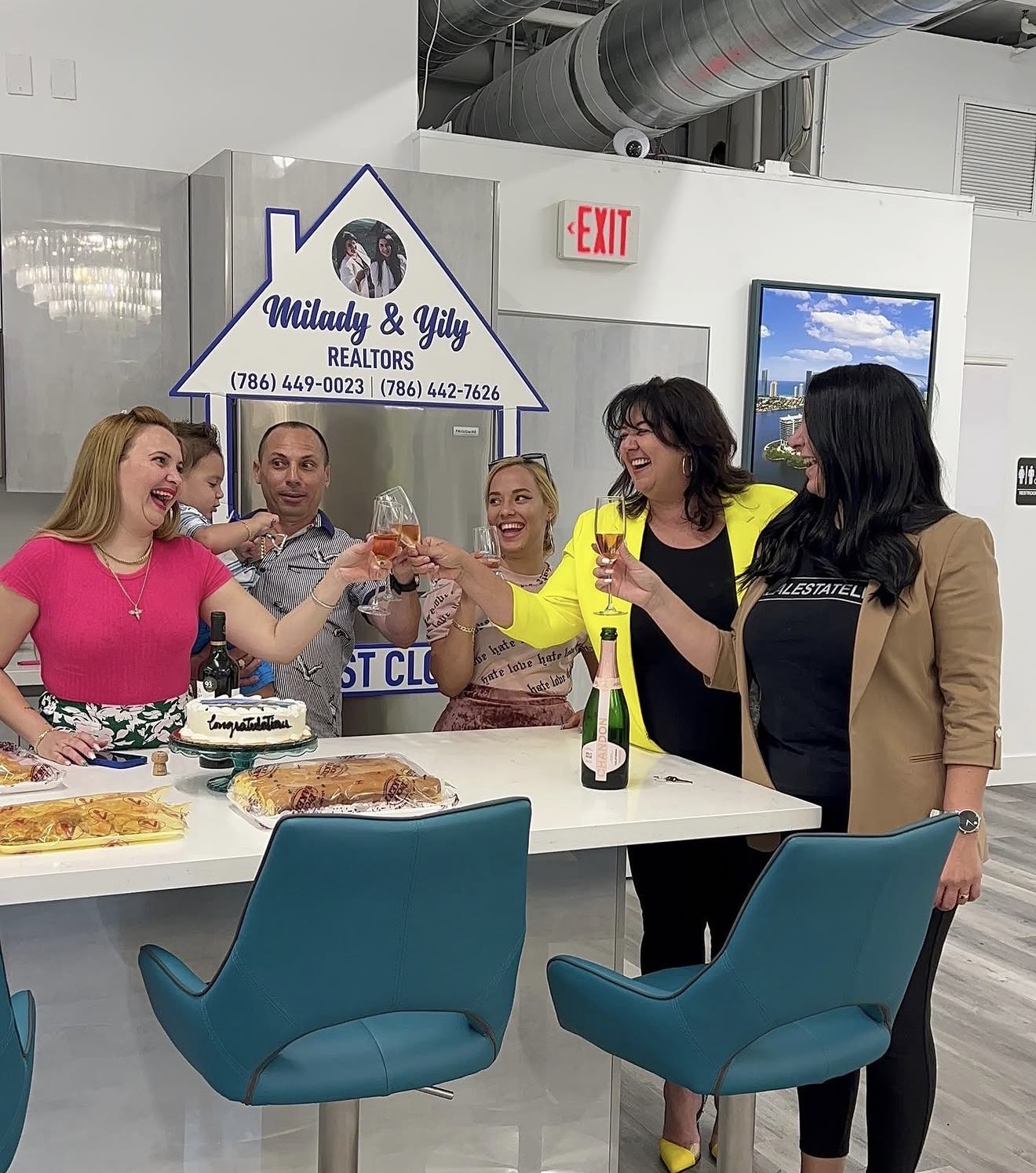 Group of women celebrating around a table with cake, smiling and clapping, in an office setting.