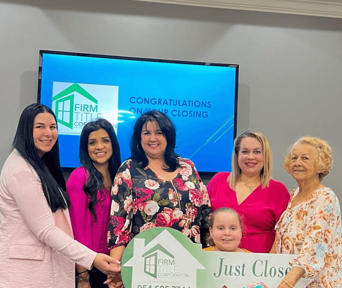Group of six women and one girl holding a large check in front of a screen with a logo and message.