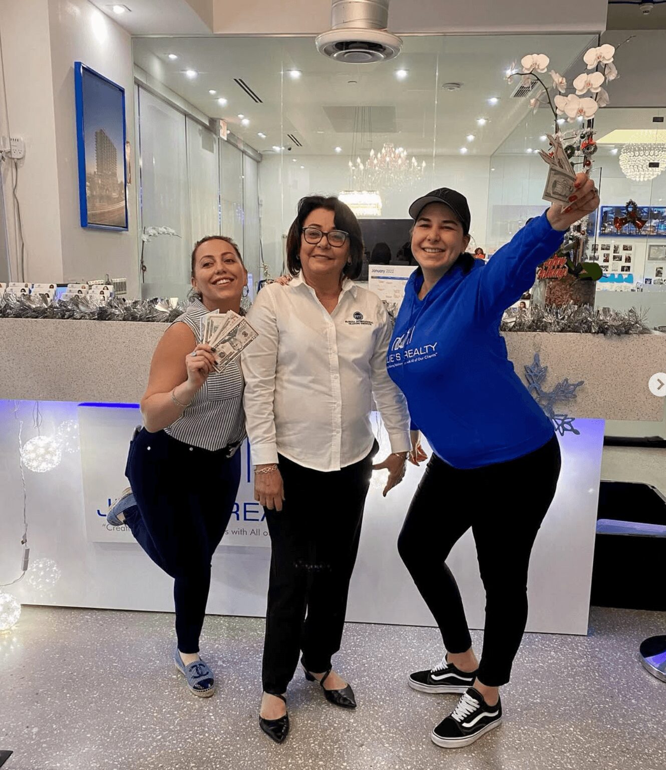 Three women standing in front of a reception desk, smiling, with one holding a bouquet of flowers and another holding money.