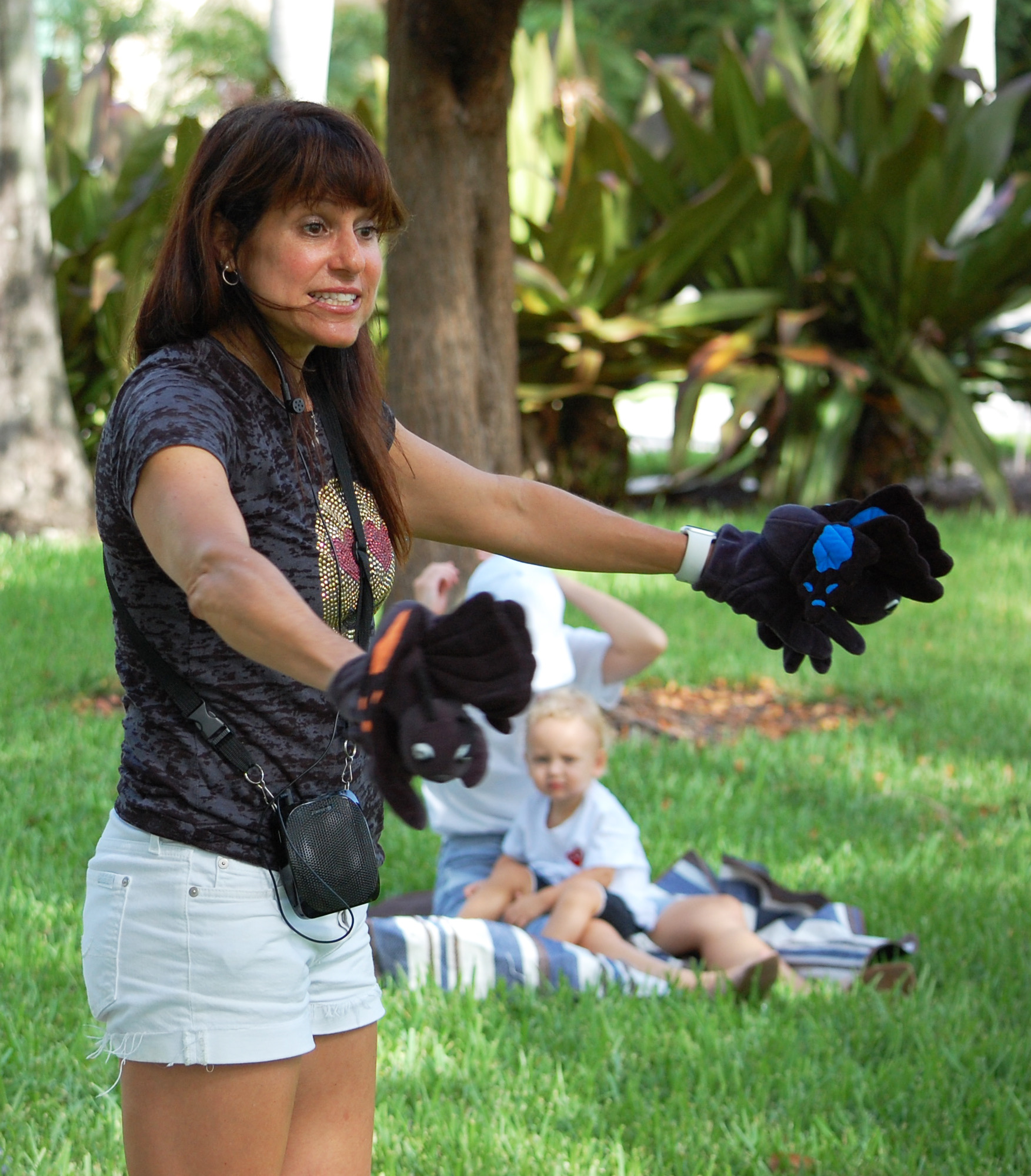 Woman holding black gloves outdoors with children sitting on grass in background.