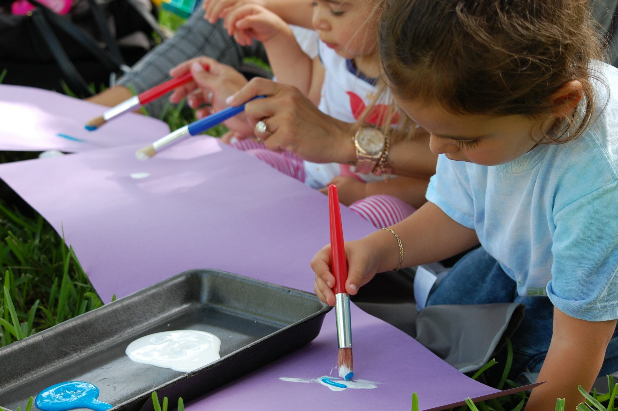 Children paint on large purple paper outdoors, using brushes and markers, with a tray of paint nearby.