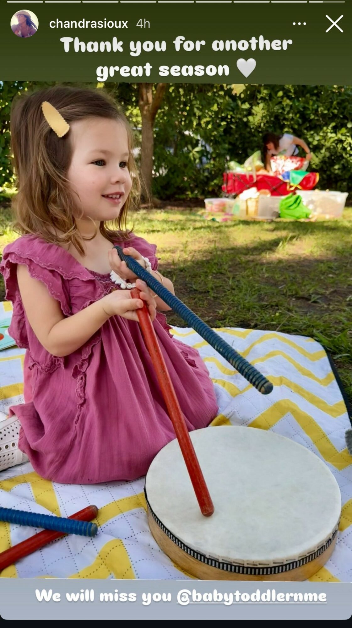 Young girl in pink dress playing a drum outdoors on a yellow and white blanket.
