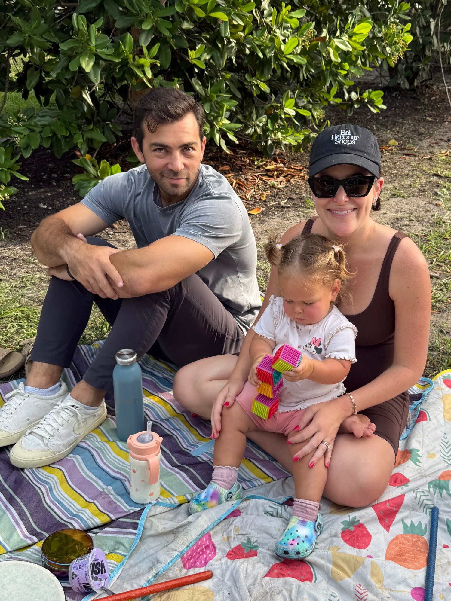 Three people sitting on a colorful blanket outdoors, with greenery in the background. A man, woman, and young girl are present.