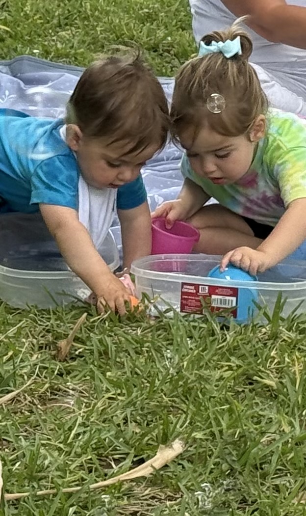Two children lying on grass, playing with a container and small objects outdoors.