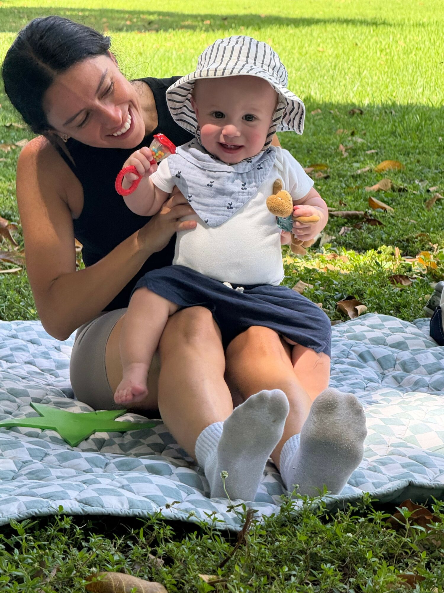 Woman and baby sitting on a blanket outdoors, woman smiling, baby wearing a hat, holding a snack, grassy background.