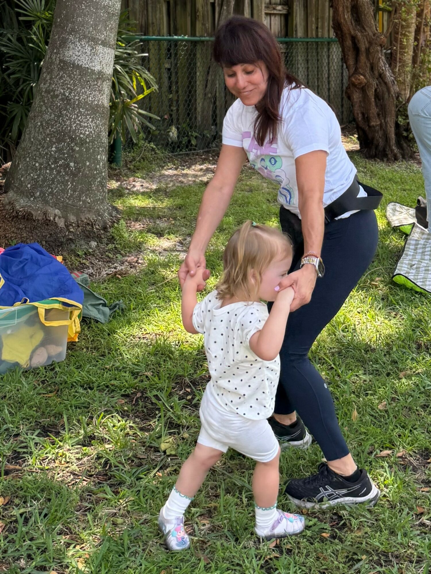 Woman and young girl dancing outdoors on grass, trees in background, woman smiling, girl in white polka dot outfit.