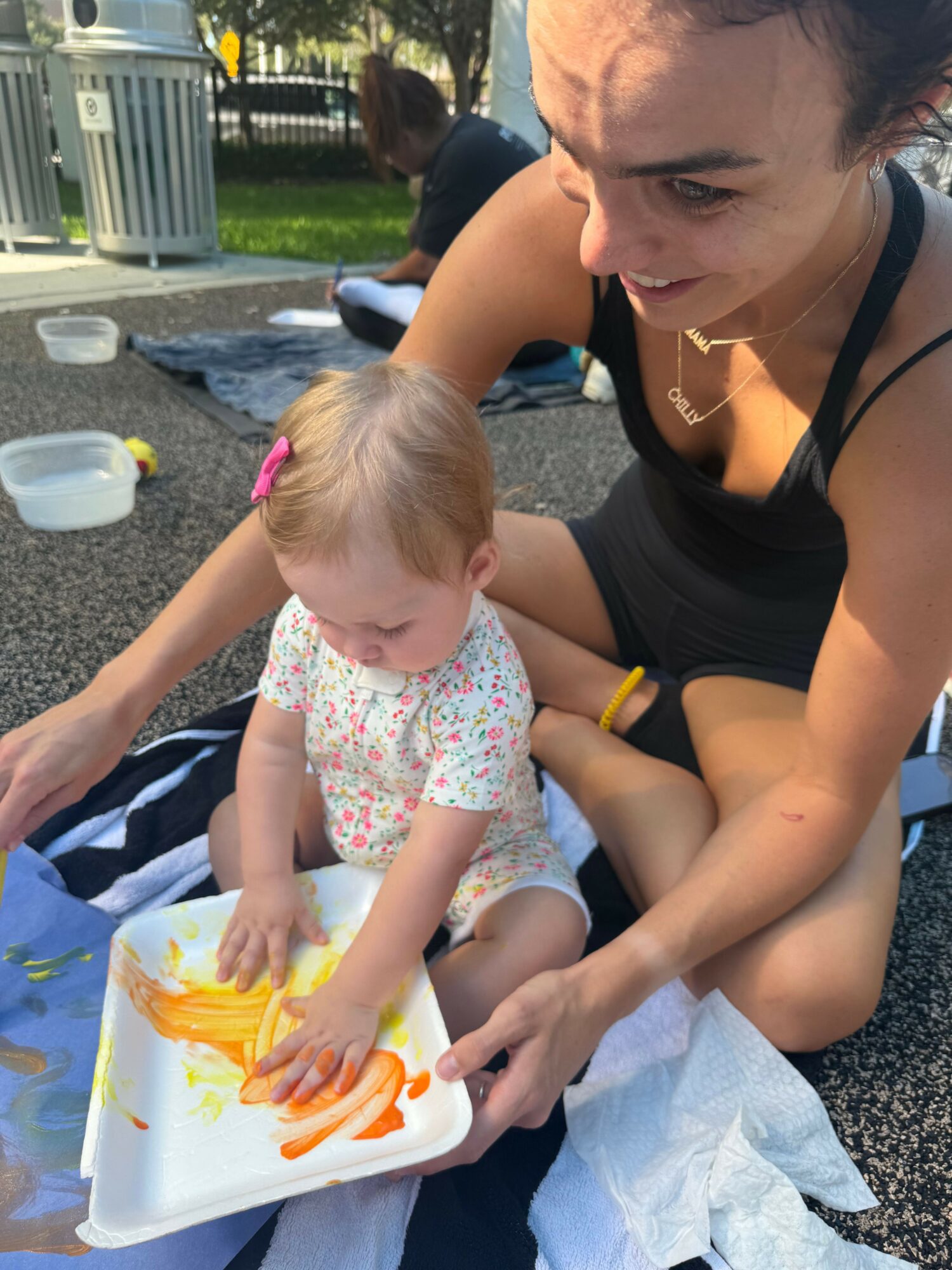 Adult and child playing with yellow and orange paint on a tray outdoors, smiling and engaging with each other.