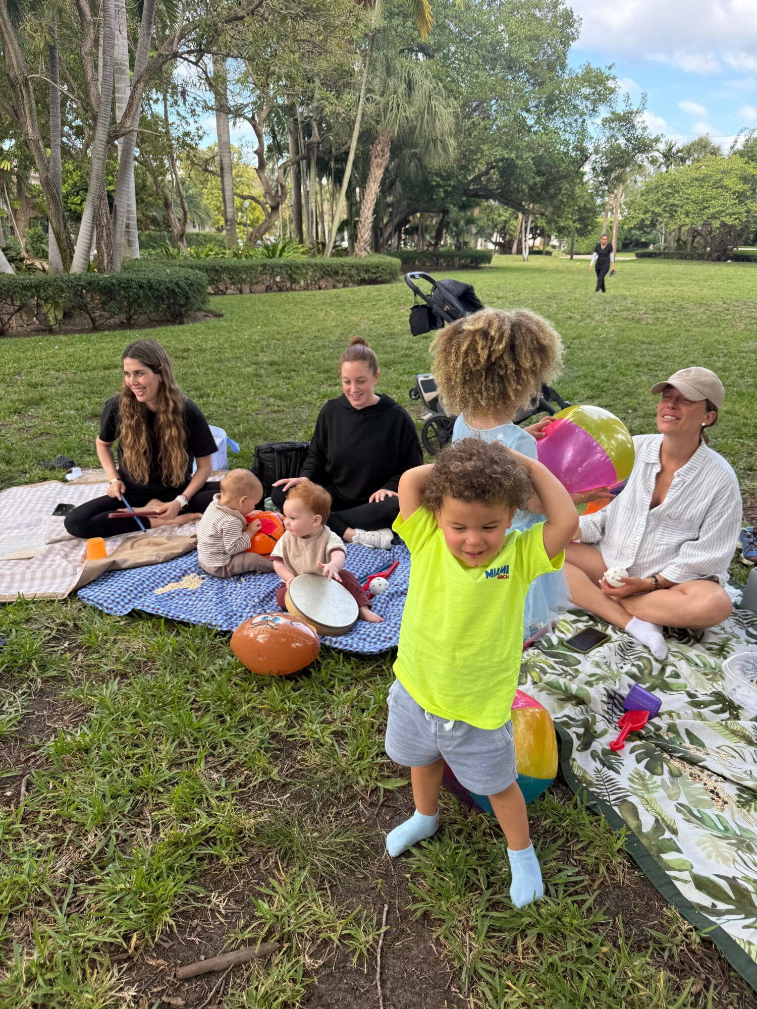 Group of people having a picnic on a grassy park with trees and a blue sky, children playing with a ball.