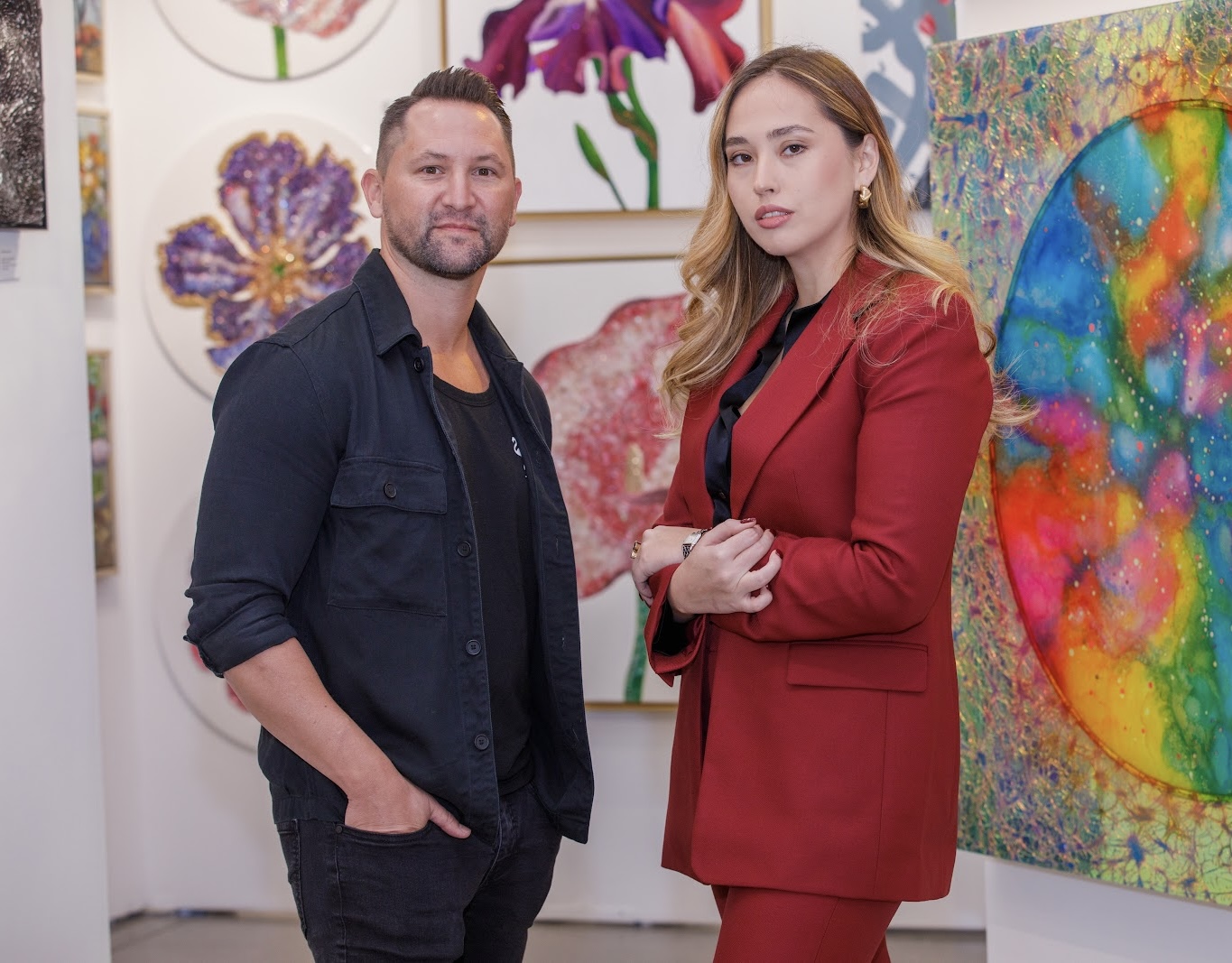 Man and woman stand in an art gallery with colorful paintings on the wall behind them.