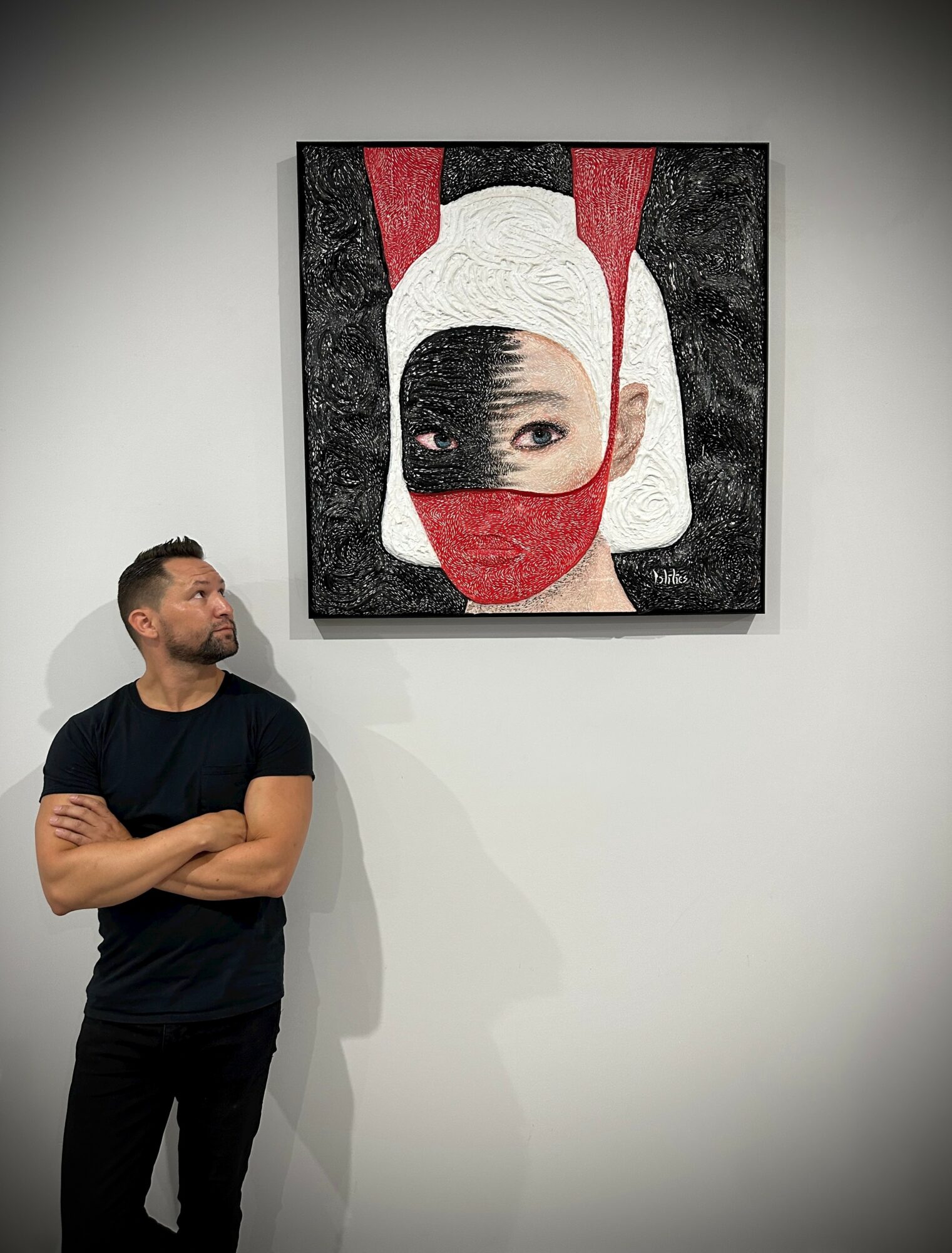 Man with arms crossed looking at artwork of a woman wearing a mask and head covering.