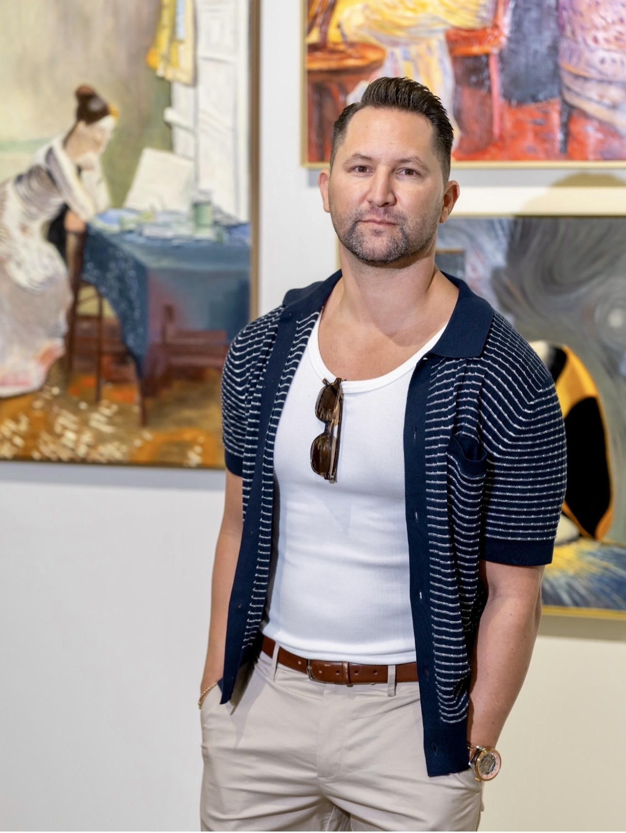Man with short dark hair, beard, wearing white t-shirt, striped navy cardigan, beige pants, sunglasses hanging, in art gallery.