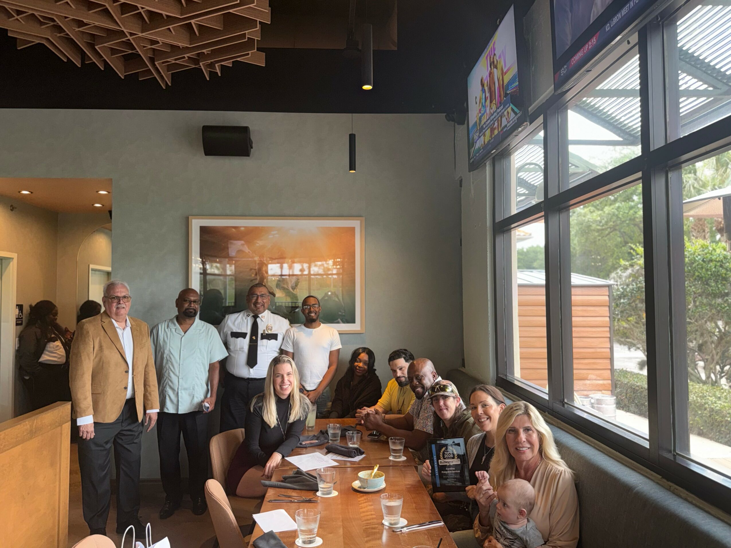 Group of people gathered around a table in a restaurant, some standing and some sitting, with large windows nearby.