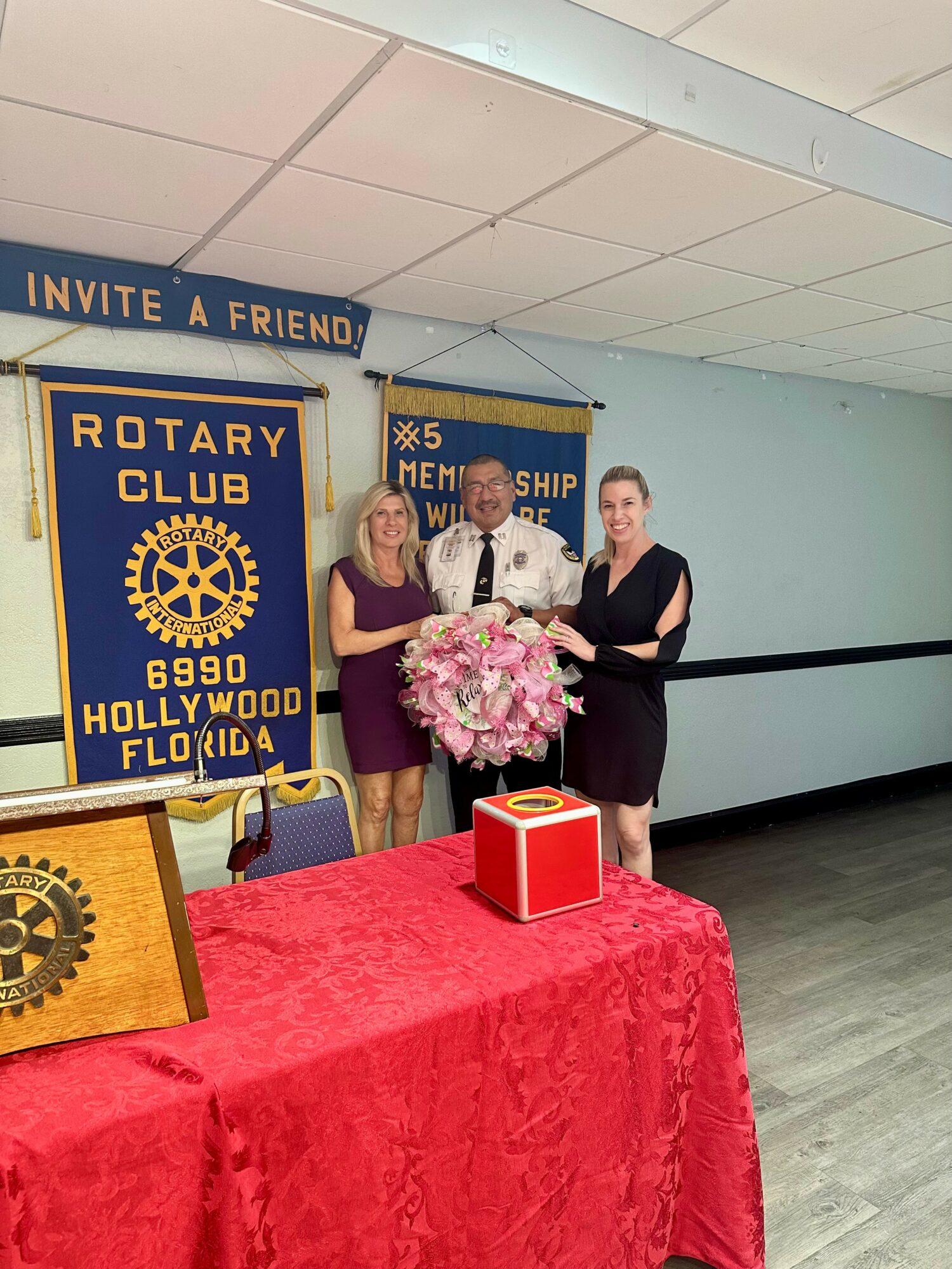 Three women standing together, one holding a large bouquet of pink flowers, in a room with banners and a table with a red cloth.