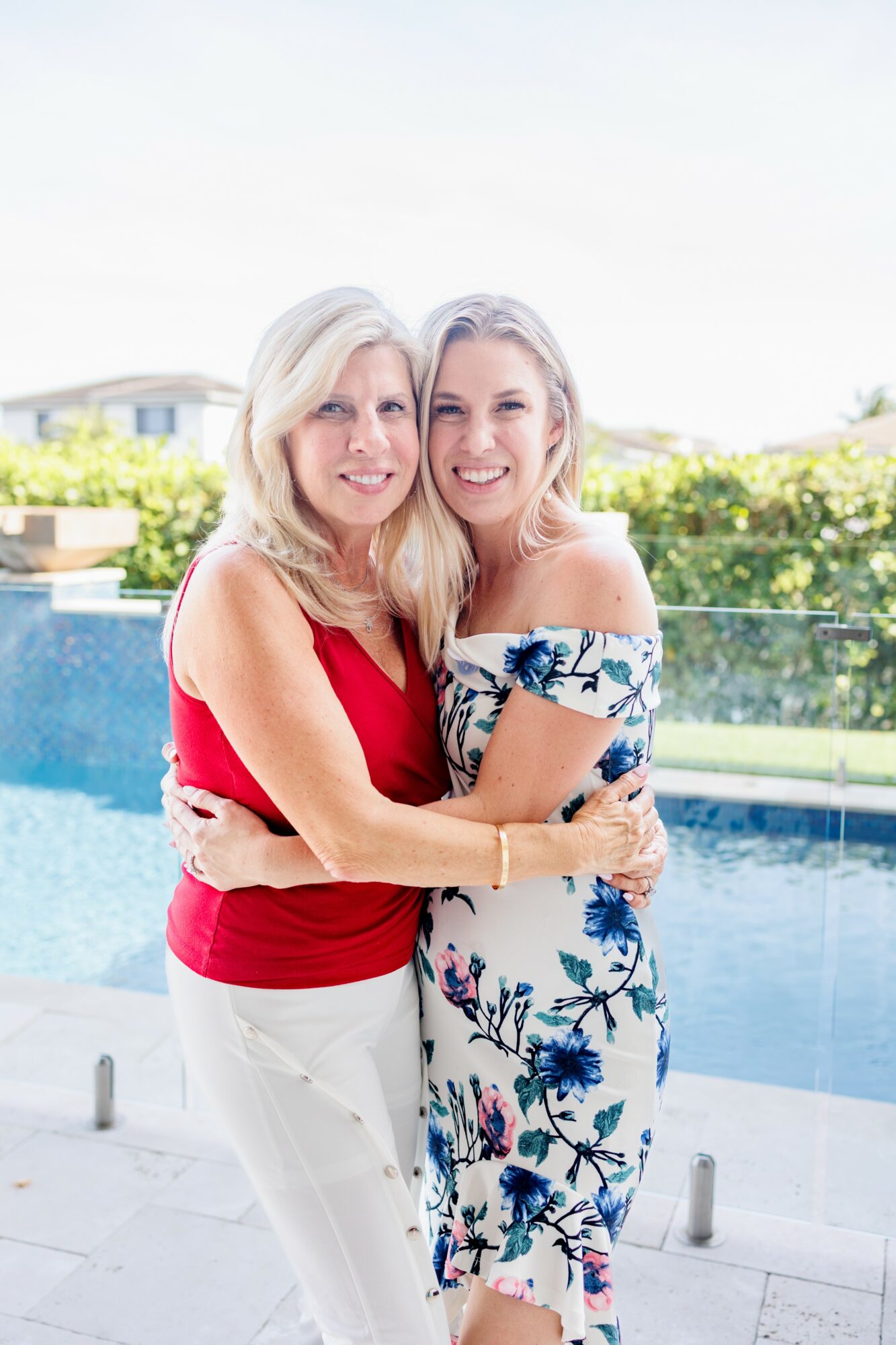 Two women with blonde hair smiling and hugging by a poolside, wearing colorful dresses.