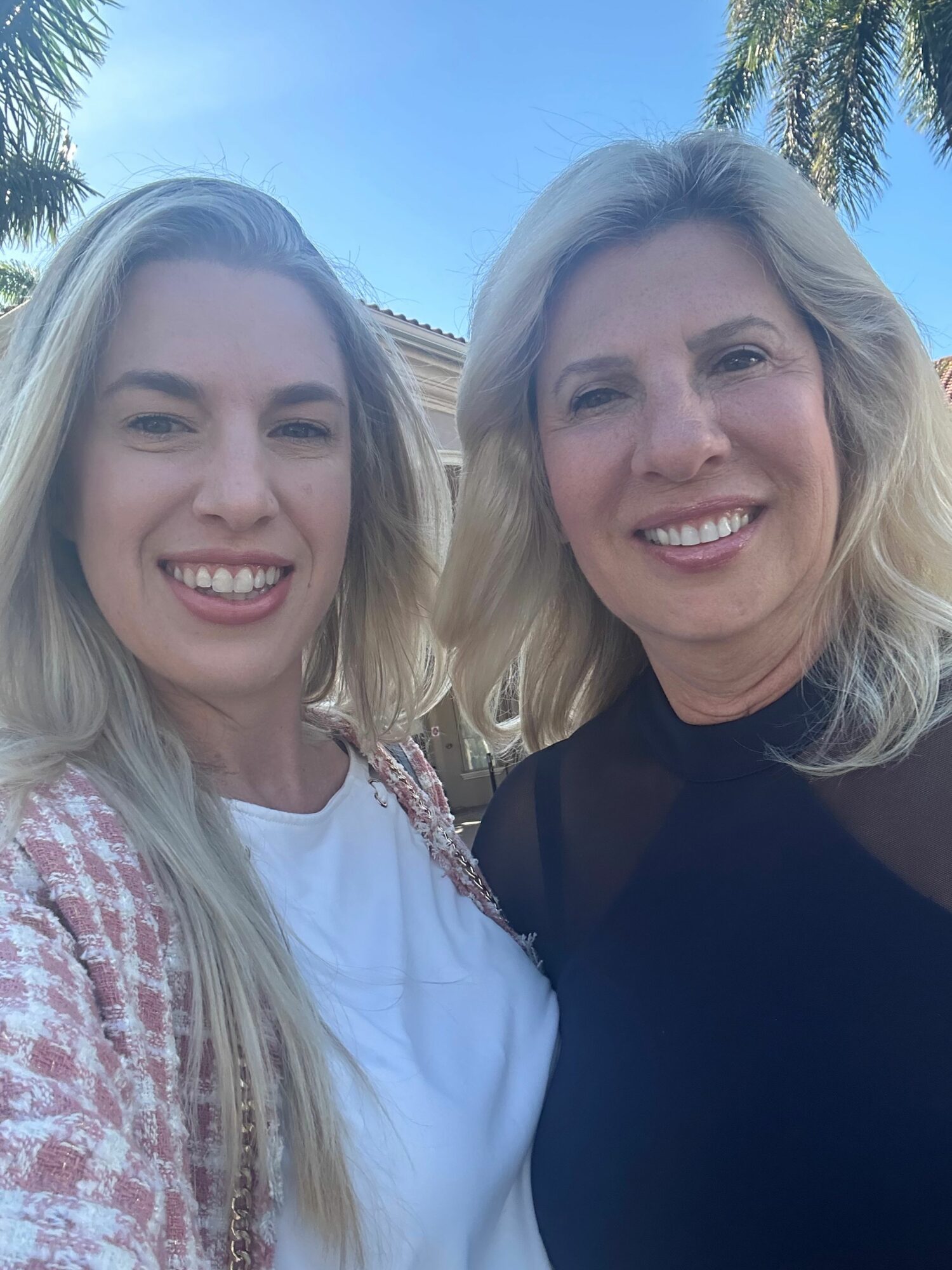 Two women smiling outdoors with trees and blue sky in background.