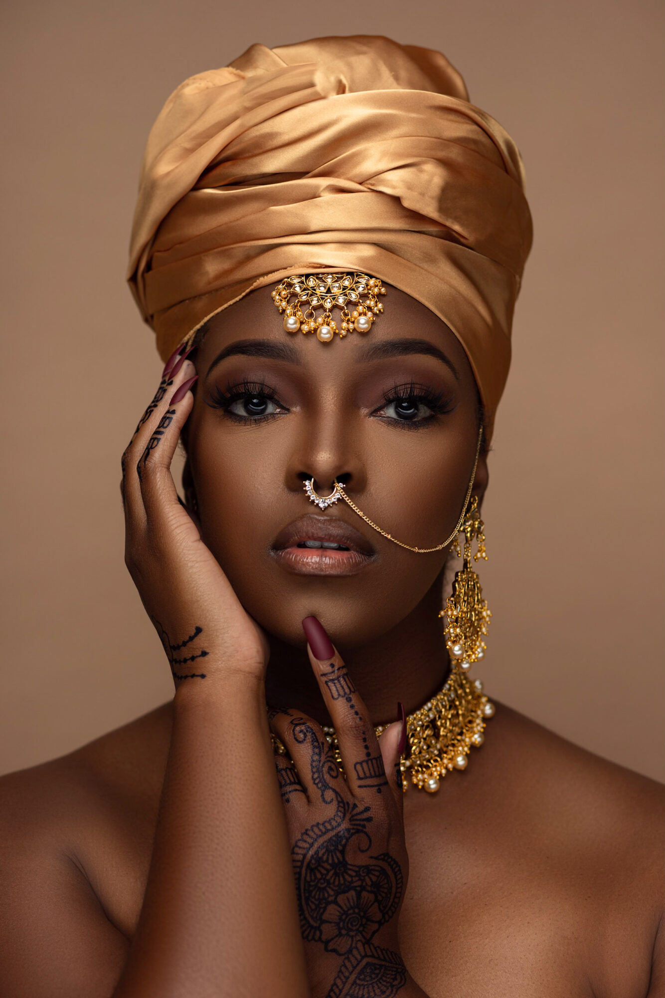 Woman with gold headwrap, jewelry, and henna tattoos, touching her face with one hand, against a plain background.