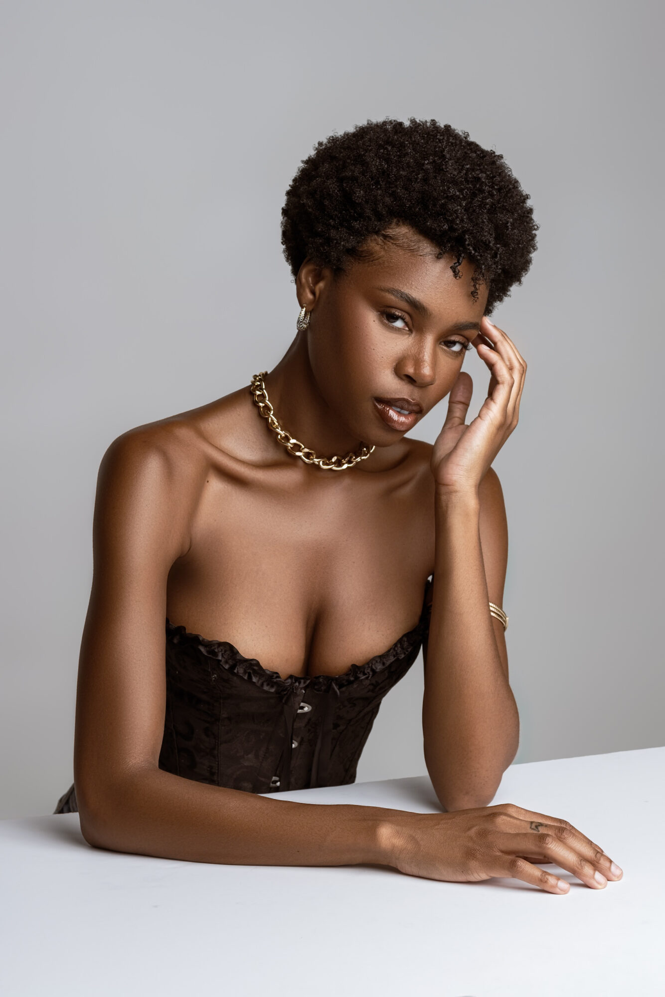 Young woman with short curly hair, wearing a strapless black top and gold jewelry, resting her head on her hand.