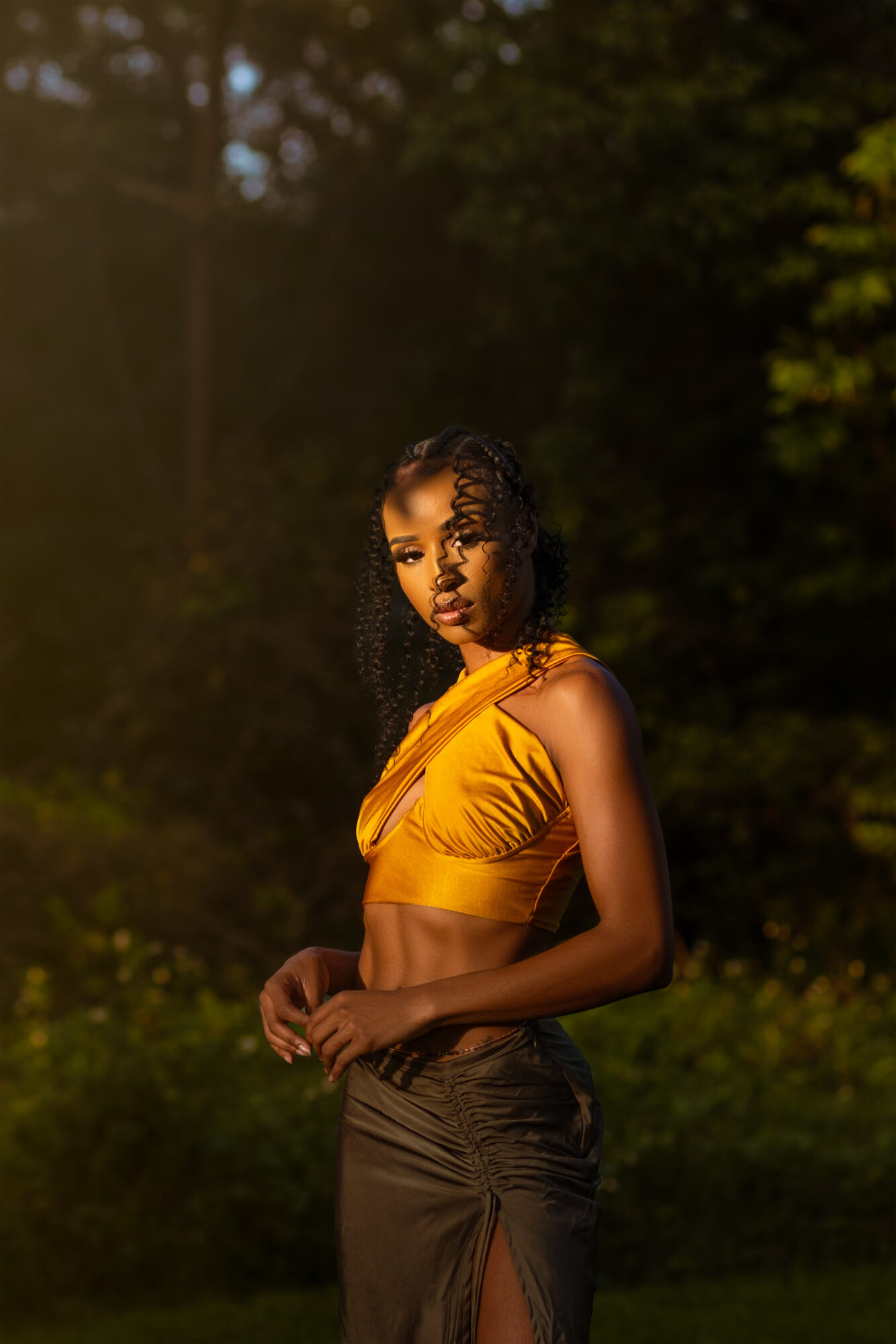 Young woman with curly hair in yellow crop top and dark skirt outdoors, trees in background.