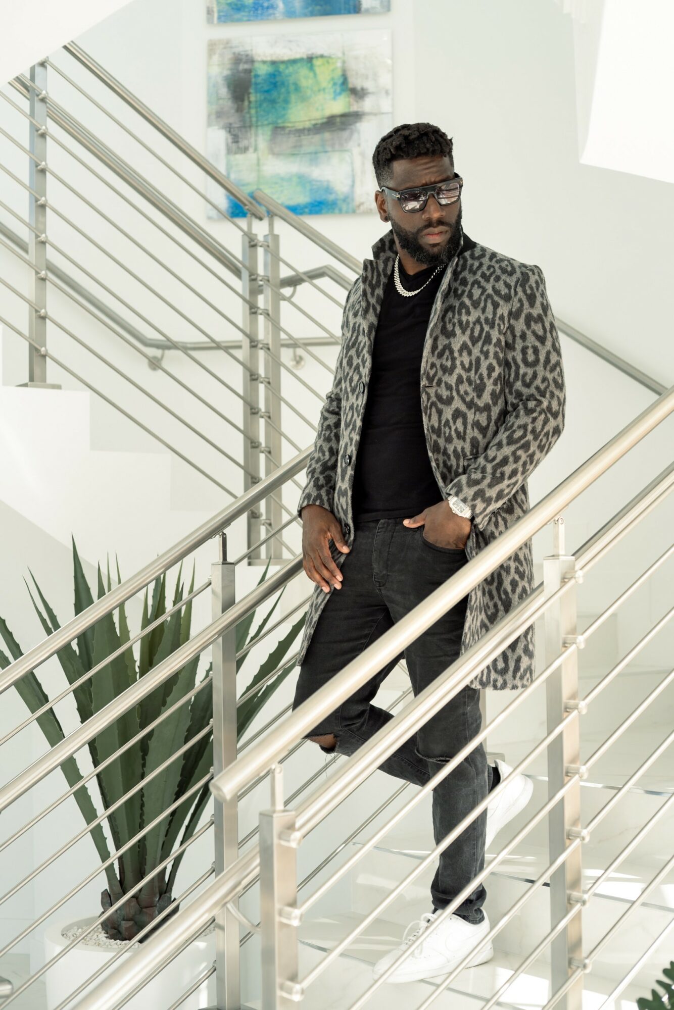 Man with glasses and a leopard print coat standing on a staircase indoors, with railing and artwork in background.