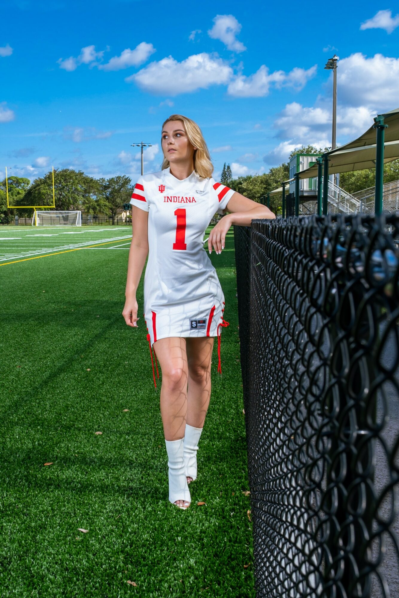 Female athlete in a white sports uniform with red accents, standing on a green field near a black fence.