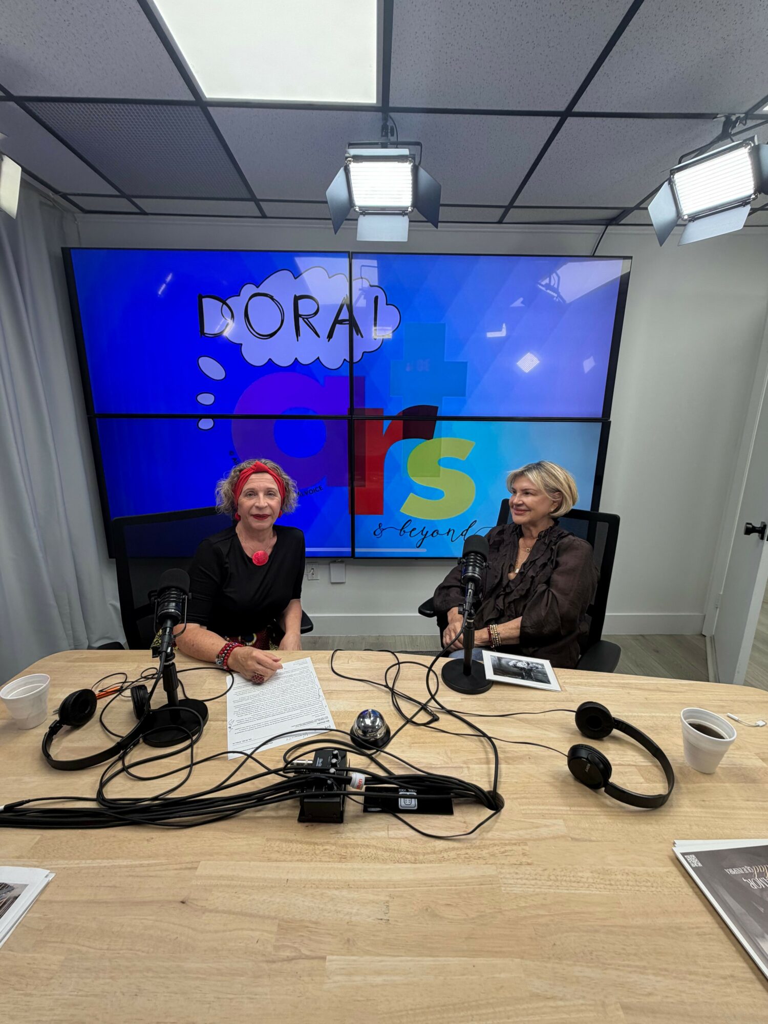 Two women sit at a table with microphones and headphones, in a room with a large screen behind them displaying colorful graphics.