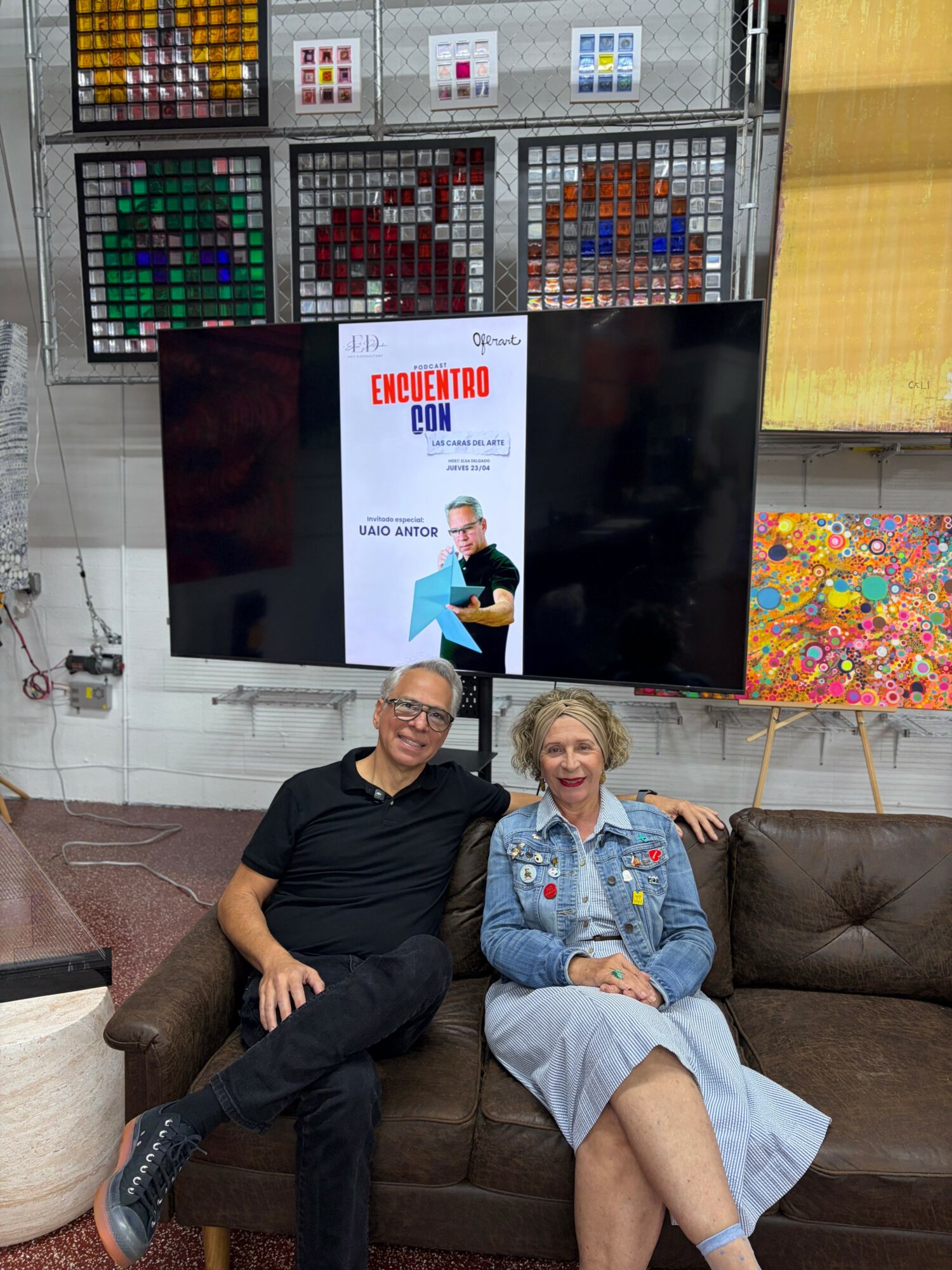 Two people sitting on a brown couch, smiling, with pixel art and a poster behind them.