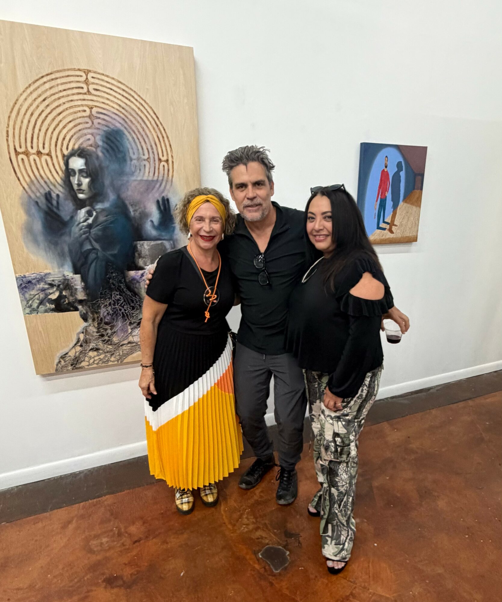 Three people stand together in an art gallery, smiling, with two artworks on the white wall behind them.