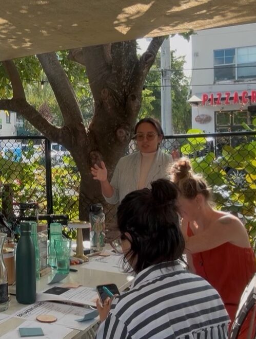 Three women sitting outdoors at a table with drinks, one standing near a tree, holding a phone, in a casual setting.
