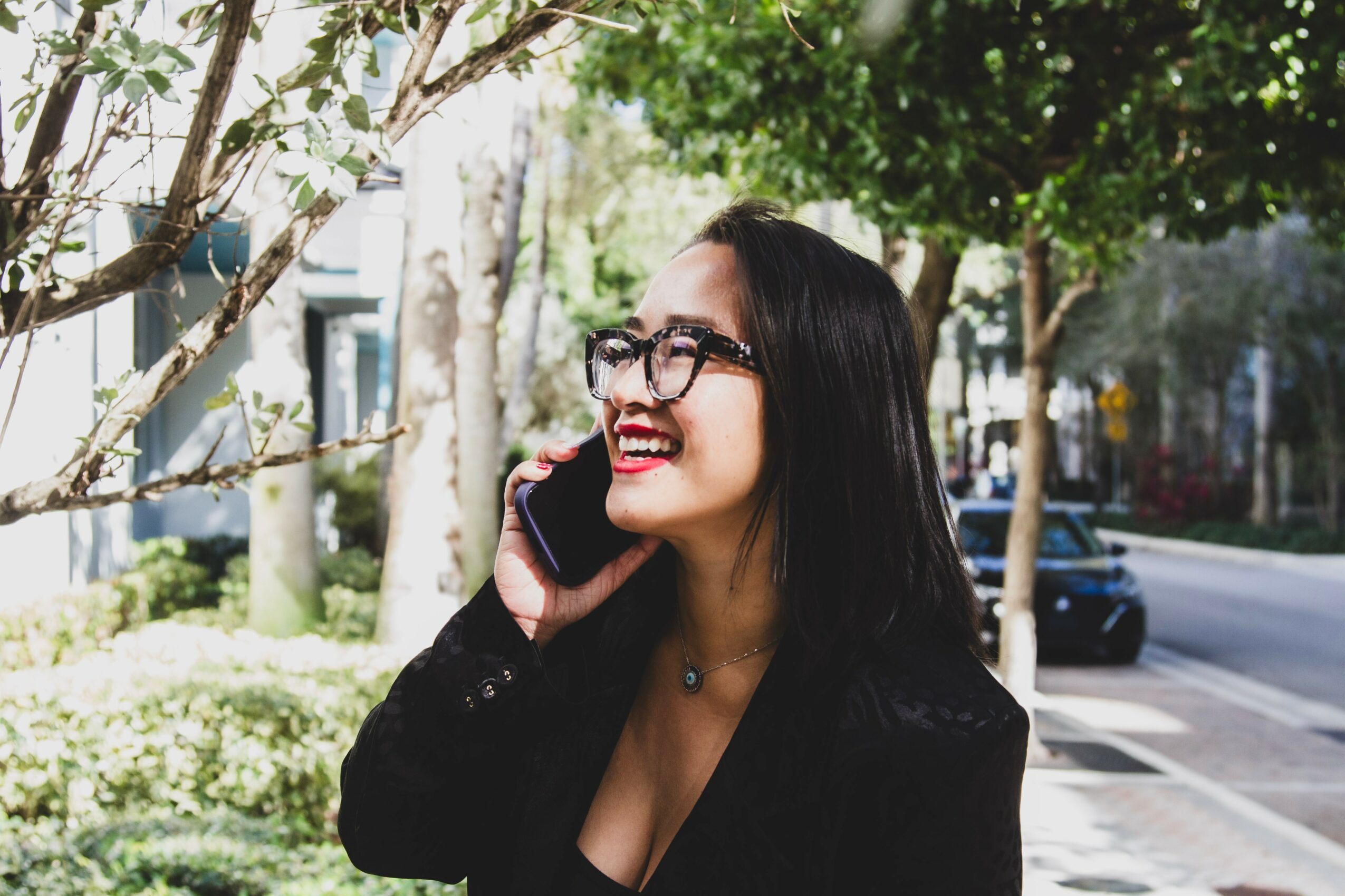 Woman with glasses talking on a mobile phone outdoors, smiling, with trees and a street in the background.