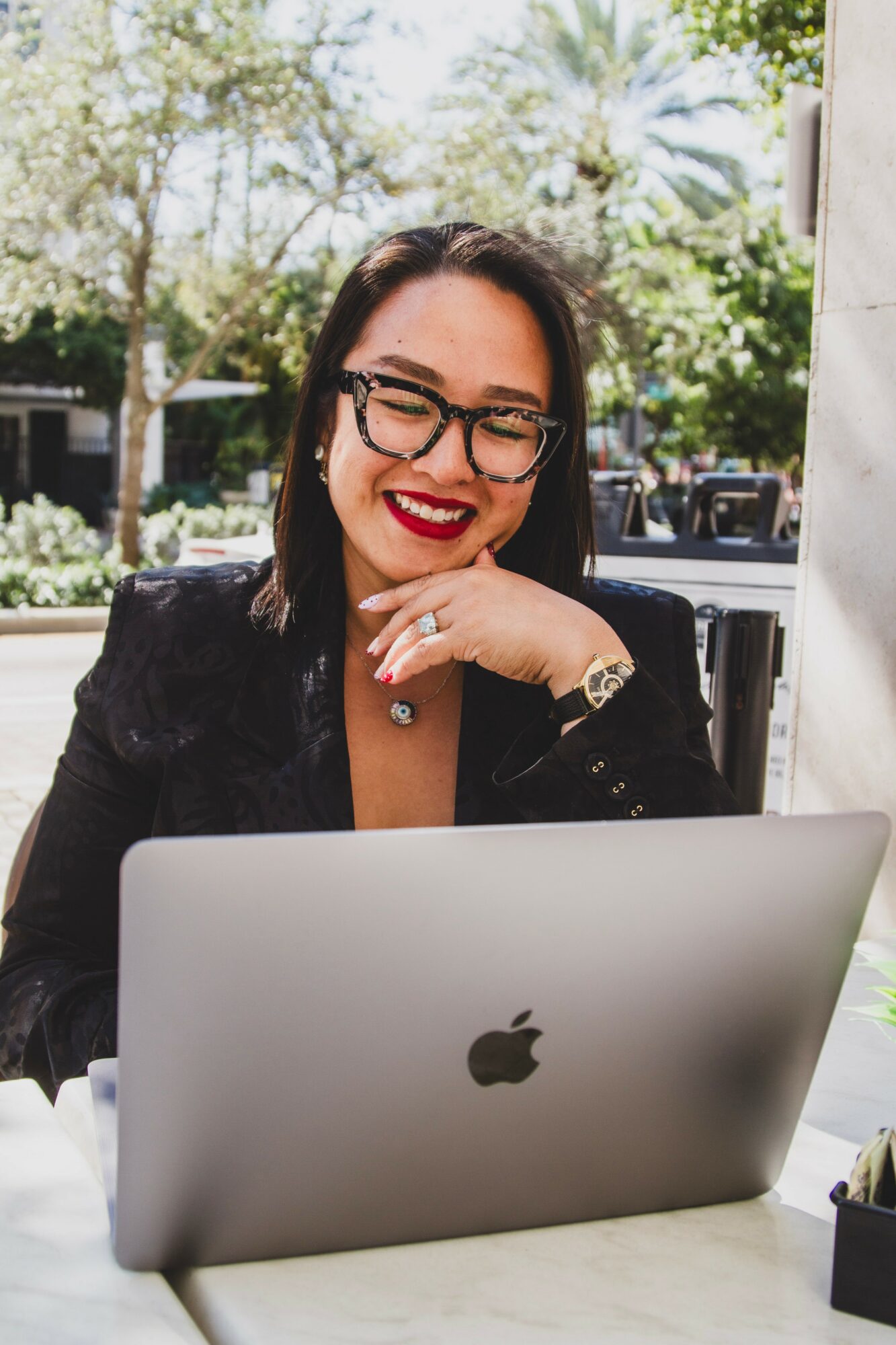 Woman with glasses smiling at a laptop outdoors, hand near chin, trees and sunlight in background.