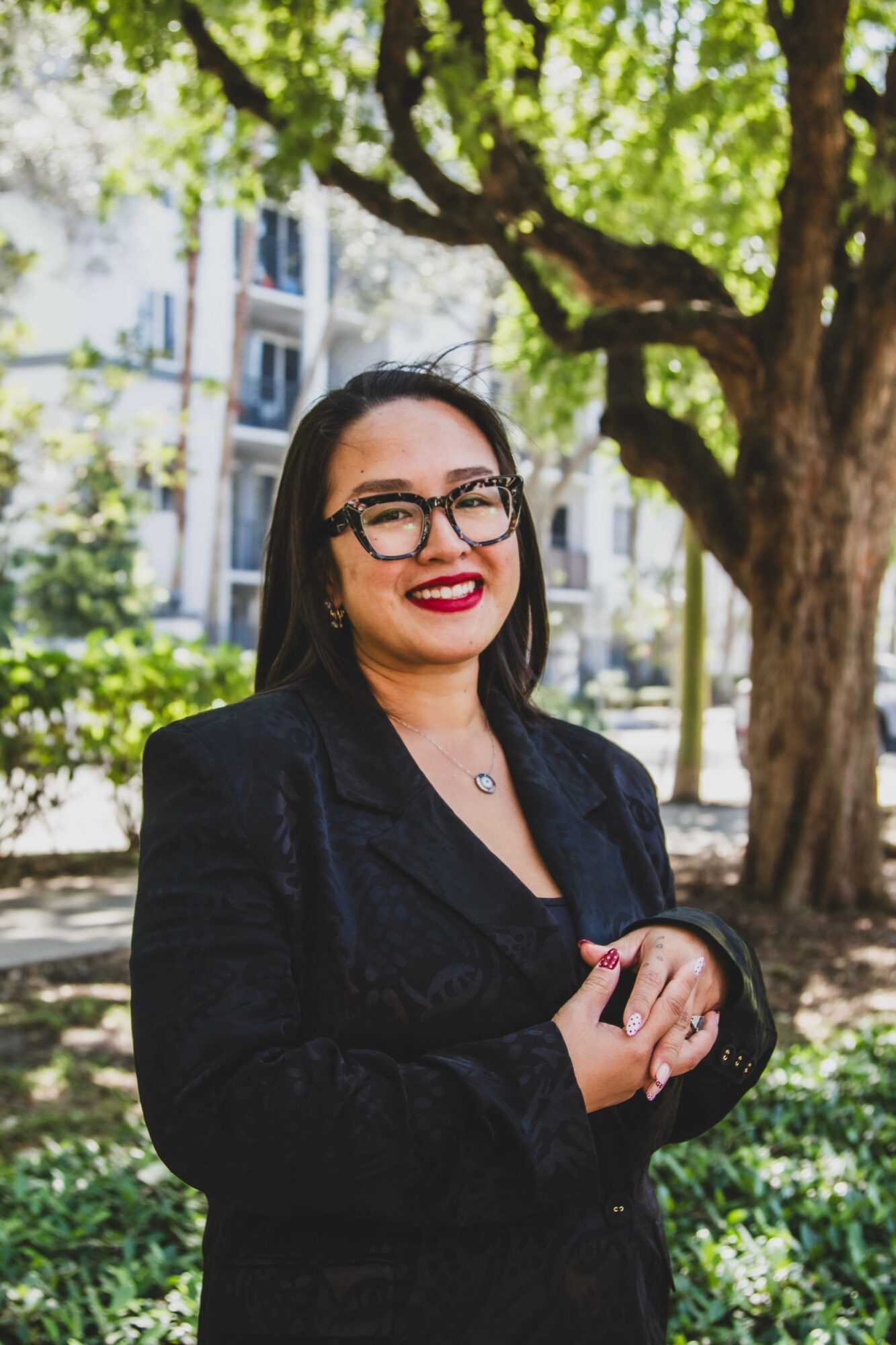 Woman with glasses and dark hair smiling outdoors, wearing a black blazer, in front of a tree and building.