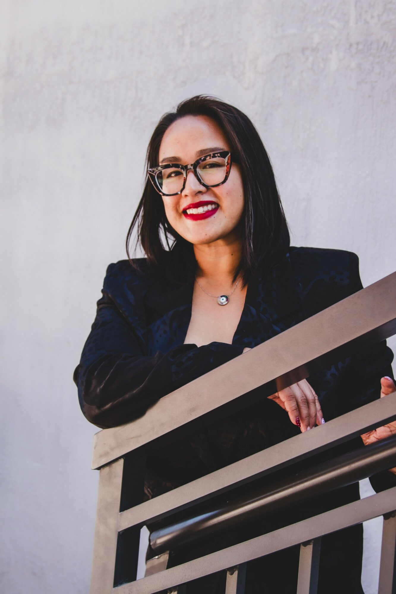 Woman with glasses smiling, wearing a dark blazer, standing near a metal railing against a plain background.