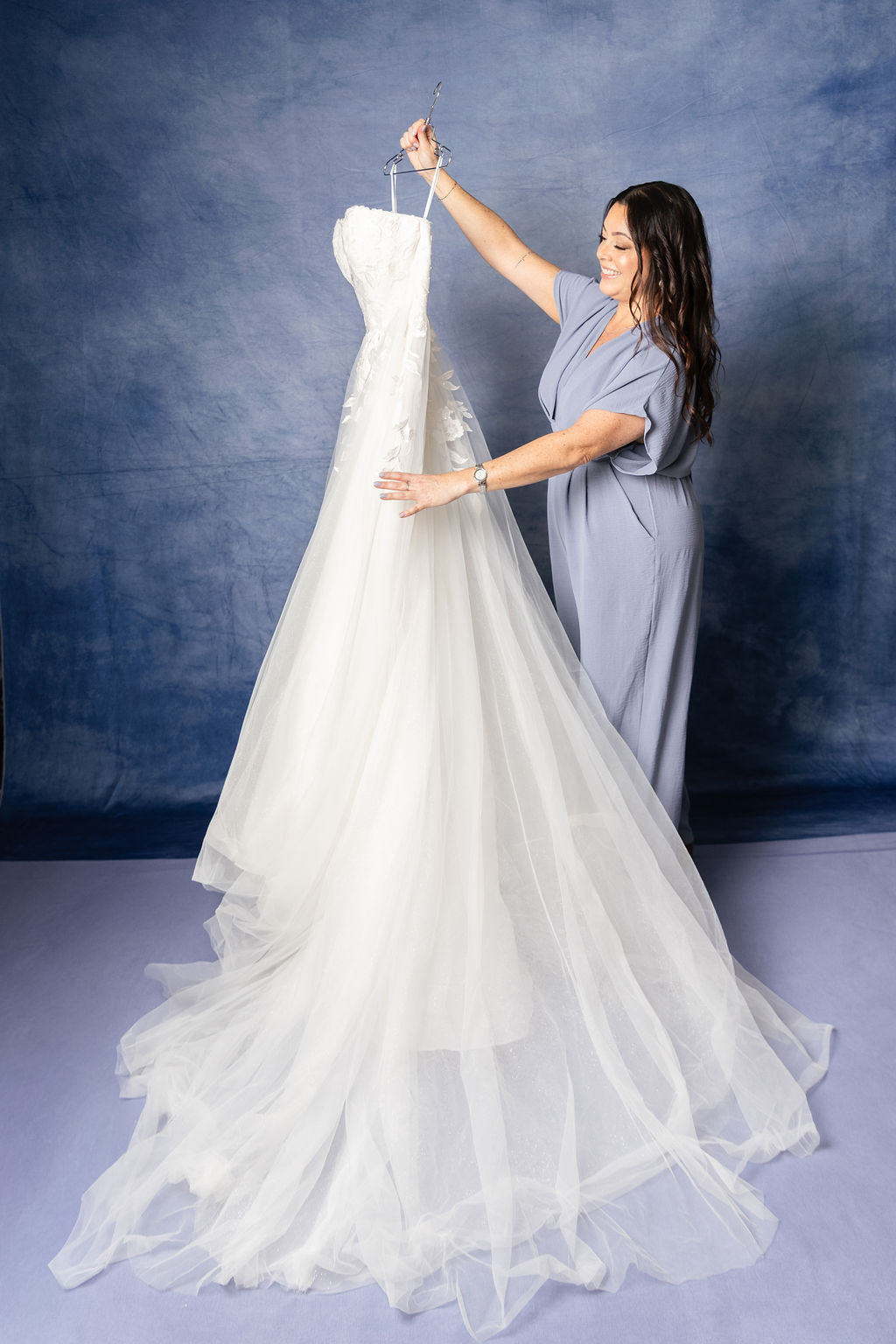 Woman in blue scrubs holding a wedding dress on a hanger, standing in front of a textured blue background.