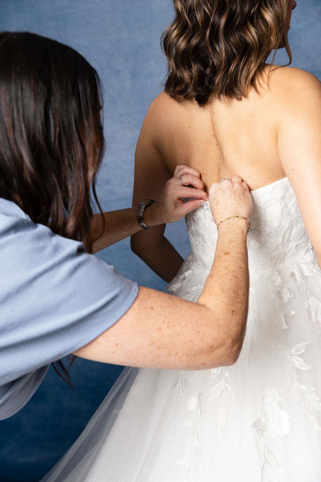 Person adjusting a wedding dress on a woman with shoulder-length hair against a blue background.