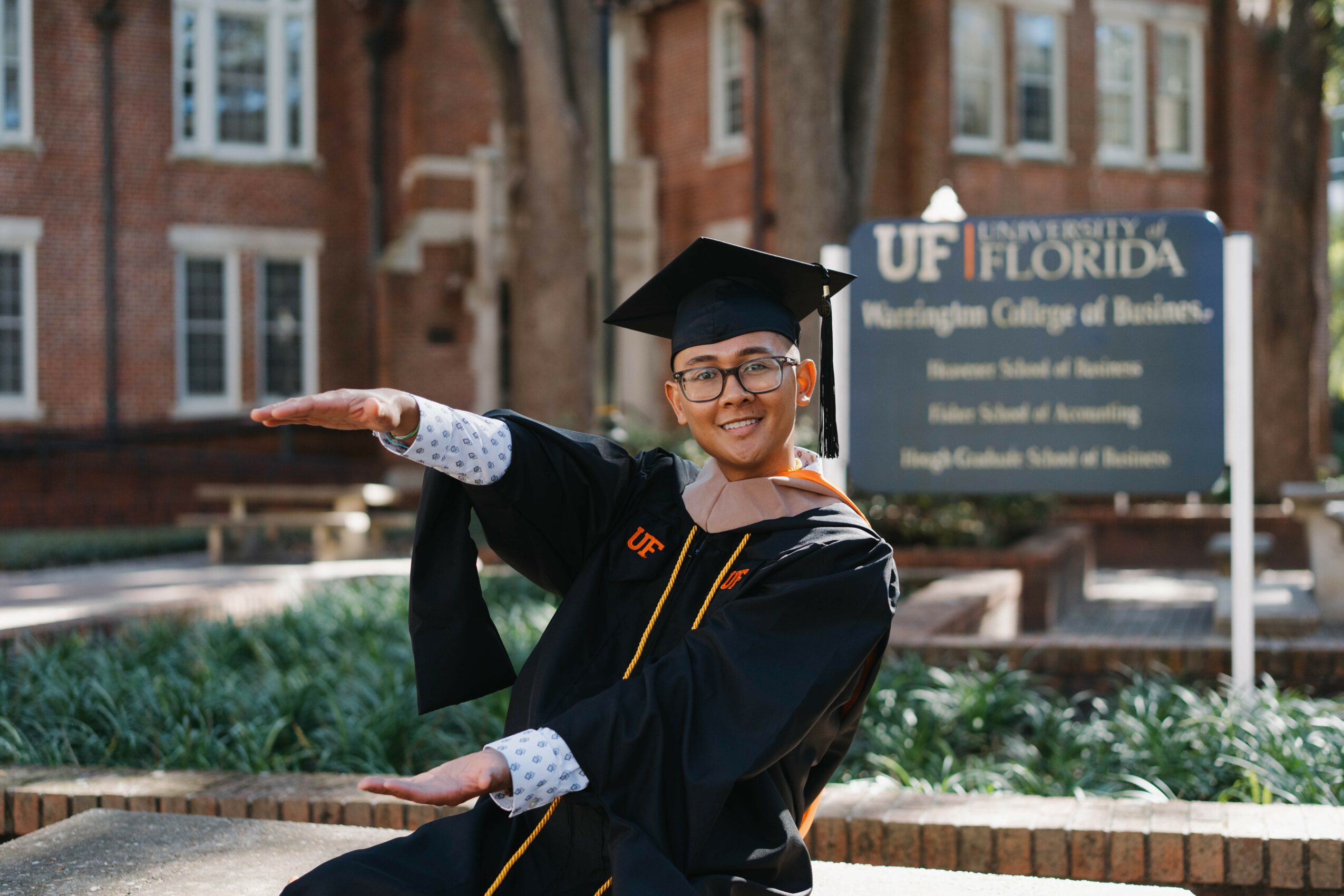 Graduate in cap and gown smiling and waving outdoors with university sign in background.