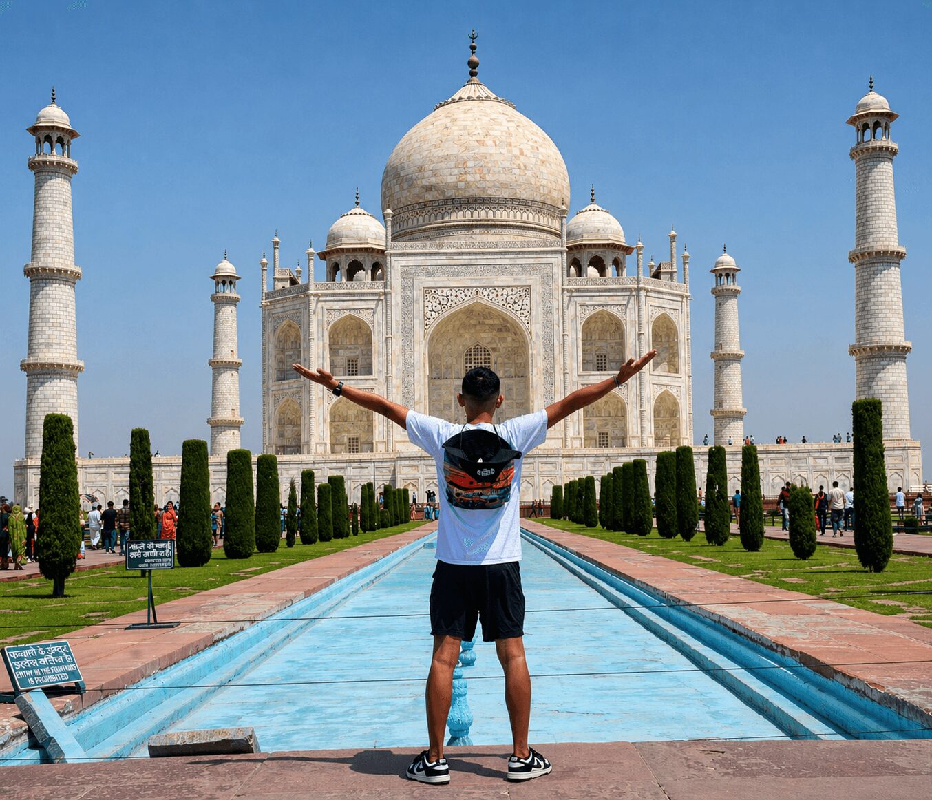 Person with arms outstretched facing the Taj Mahal, standing on a pathway with water features and greenery.