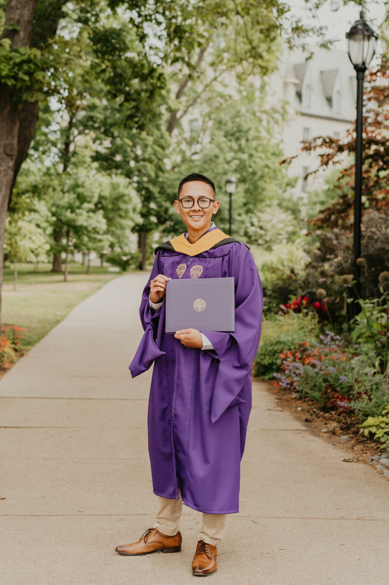 Graduate in purple gown holding a laptop, standing on a park pathway with trees and flowers.