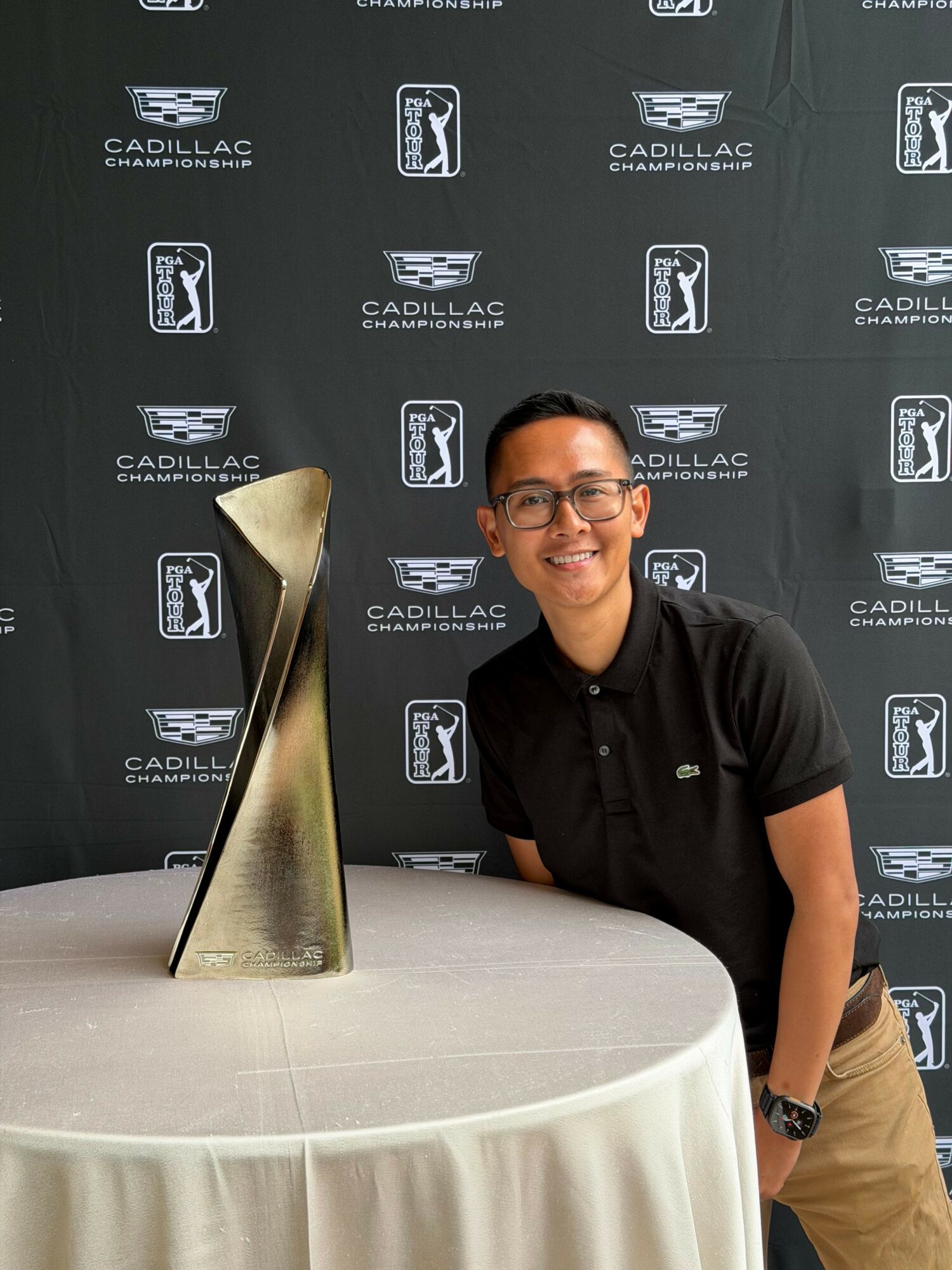 Person smiling, wearing glasses and a black polo shirt, sitting next to a tall trophy on a round table.
